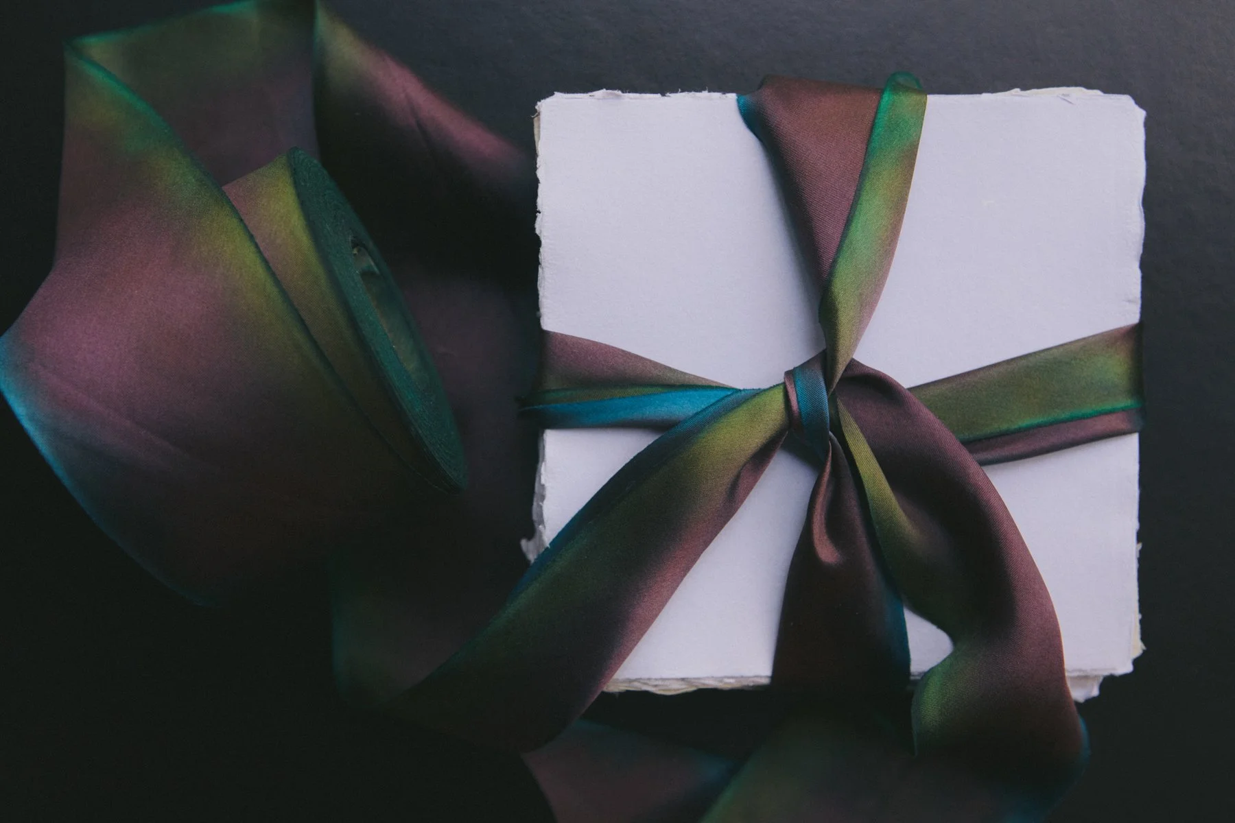 A white gift box with a glossy, rainbow-colored satin ribbon tied in a bow on top, and a matching smaller box beside it, all set against a dark background.