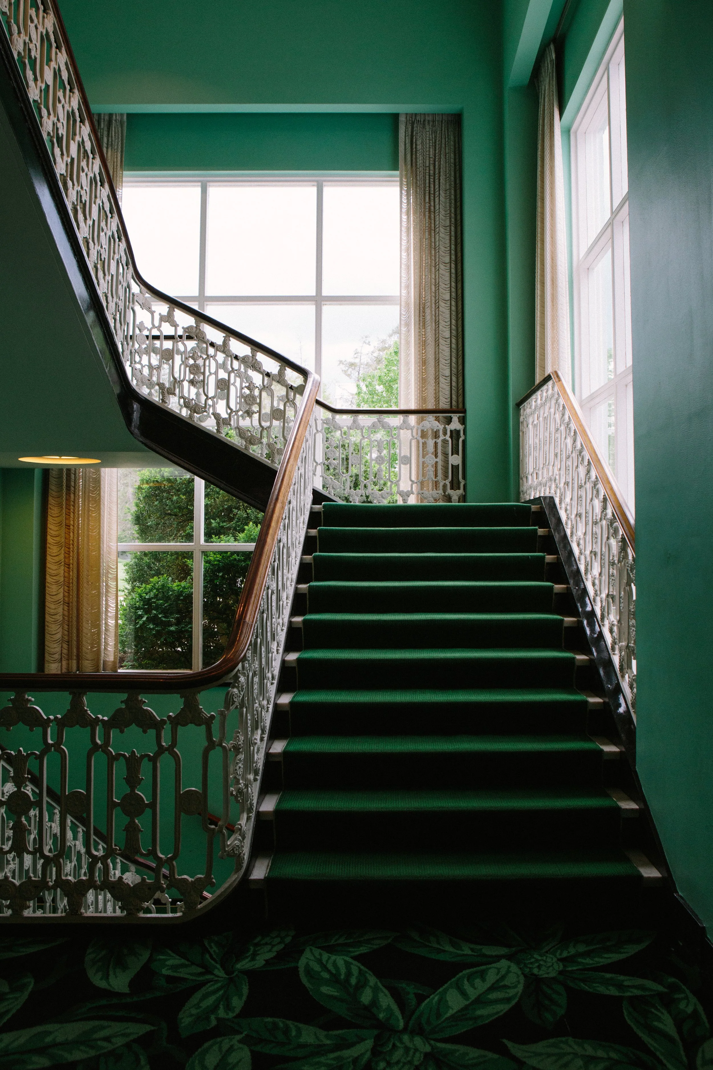 Interior view of a staircase with green steps and ornate white railing, large window letting in natural light, green walls, and floral patterned carpet on the floor.