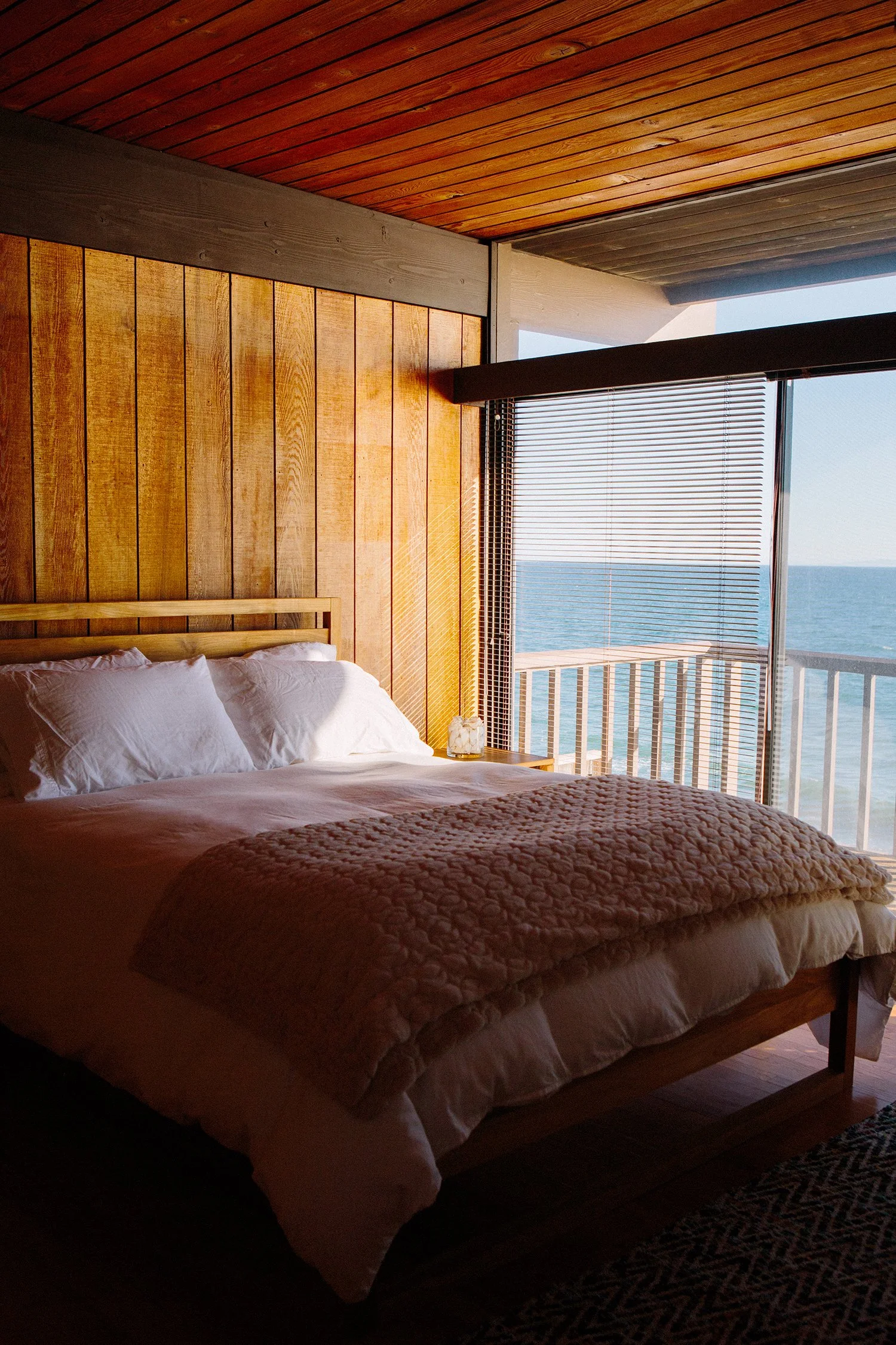 A 1970's modern bedroom with wooden walls and ceiling, a bed with white pillows and a quilted blanket, and a large window with blinds showing a view of the ocean in Malibu.