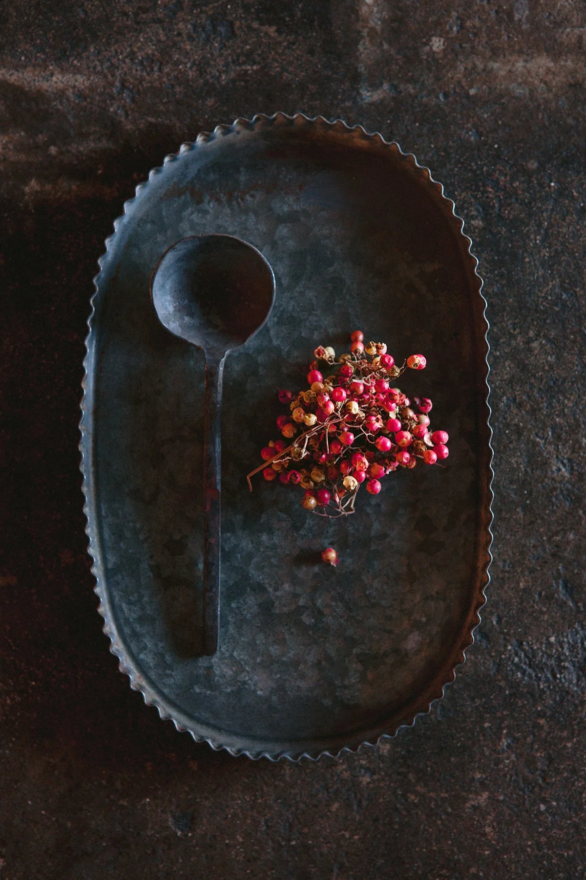 A hand-wrought iron spoon sits on a vintage metal tray next to a cluster of pink peppercorns on a dark, textured surface.