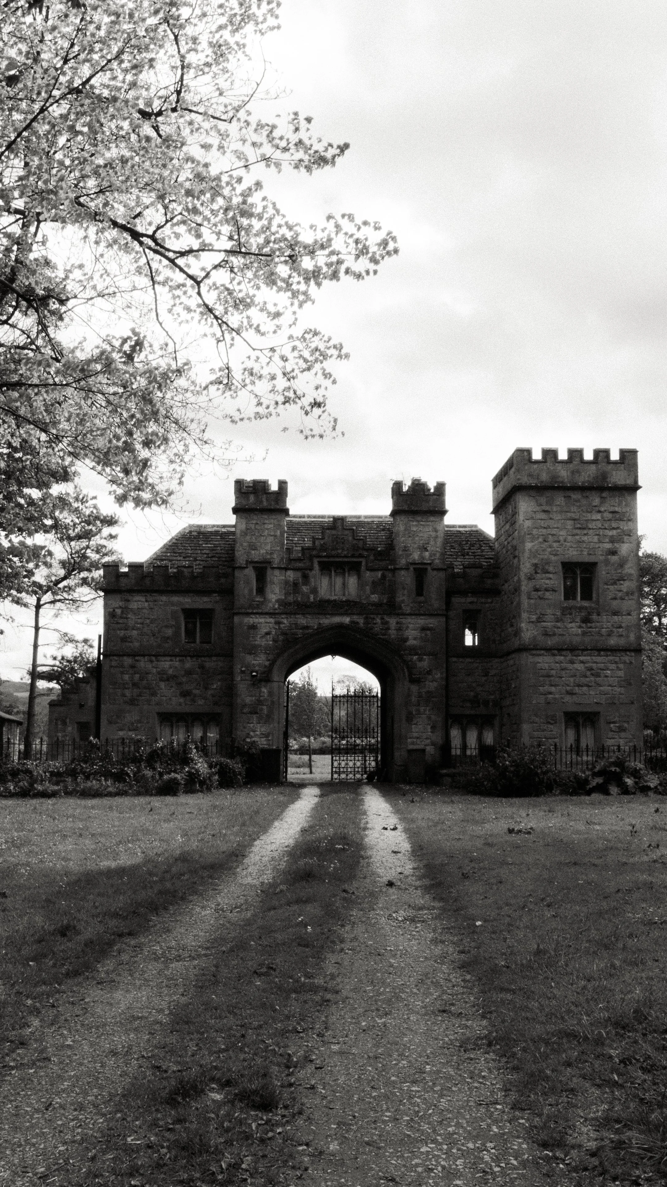 Black and white photo of a castle-like stone gate with two towers and a drawbridge in front, surrounded by trees and foliage.