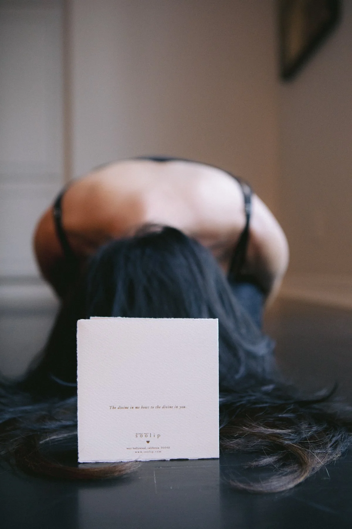 A woman with long black hair bending forward, wearing a black tank top with thin straps, with her head resting on a surface. In front of her, a white card with small text and a website address is visible. The background is neutral and out of focus.