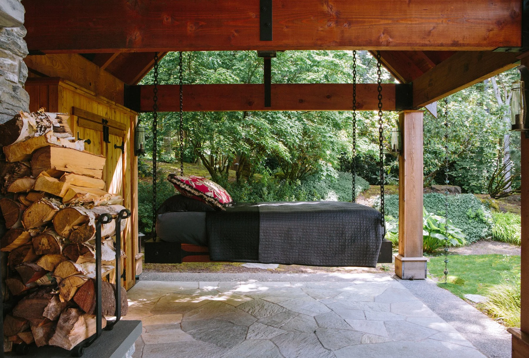 Covered outdoor patio with a hanging bed and a woodpile, surrounded by greenery. Garden Design by Bill Noland. Photographed at a private estate in Seattle, Washington.