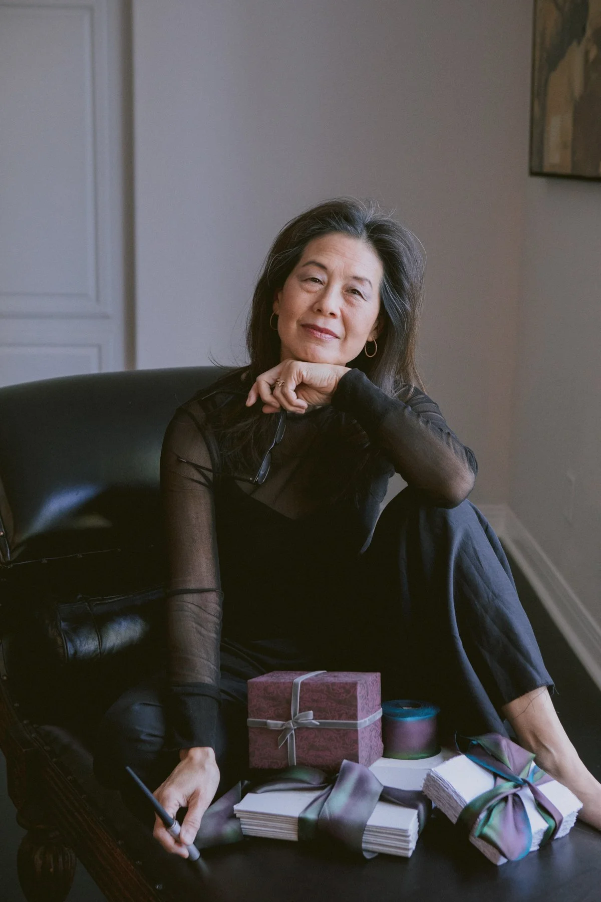 A woman sitting on a black leather chair, smiling with her chin resting on her hand, surrounded by wrapped gifts and a pen, in a room with a grey wall and a piece of artwork.
