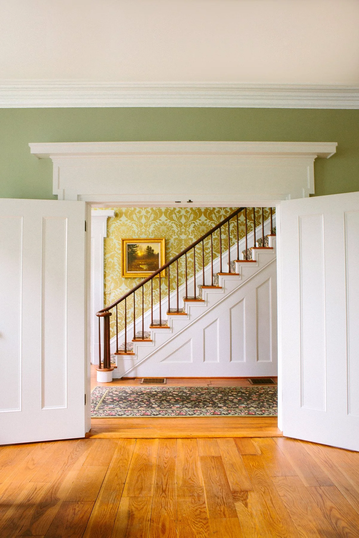View through open double white doors into a living space with a stairway, yellow Damask wallpaper, a framed landscape painting, and a patterned area rug on hardwood flooring at a private estate in Lancaster Ohio.