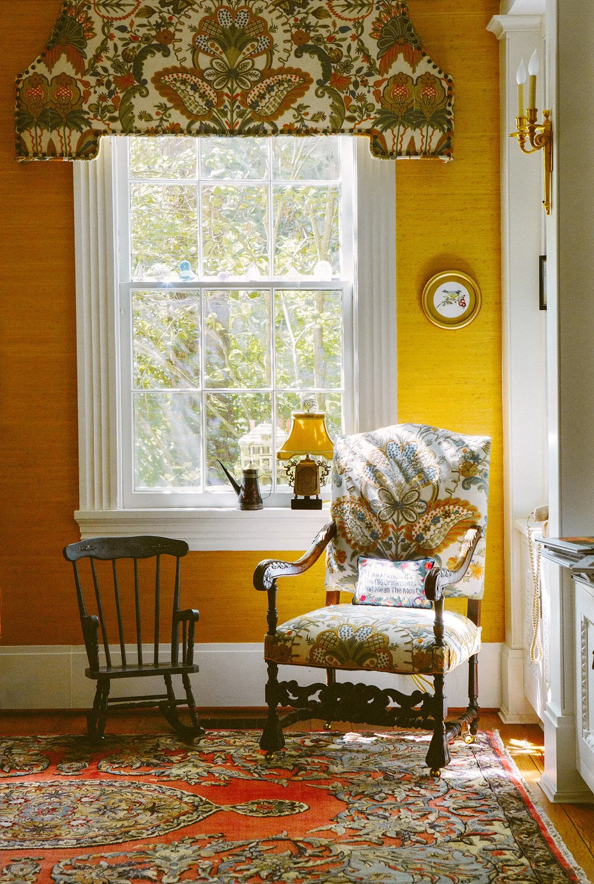 Corner of a library at a private estate in Lancaster Ohio. There is a large window, and sun streaming onto an antique chair with a floral pattern that matches the window valance and sits next to a small black rocking chair. There is a vintage rug.