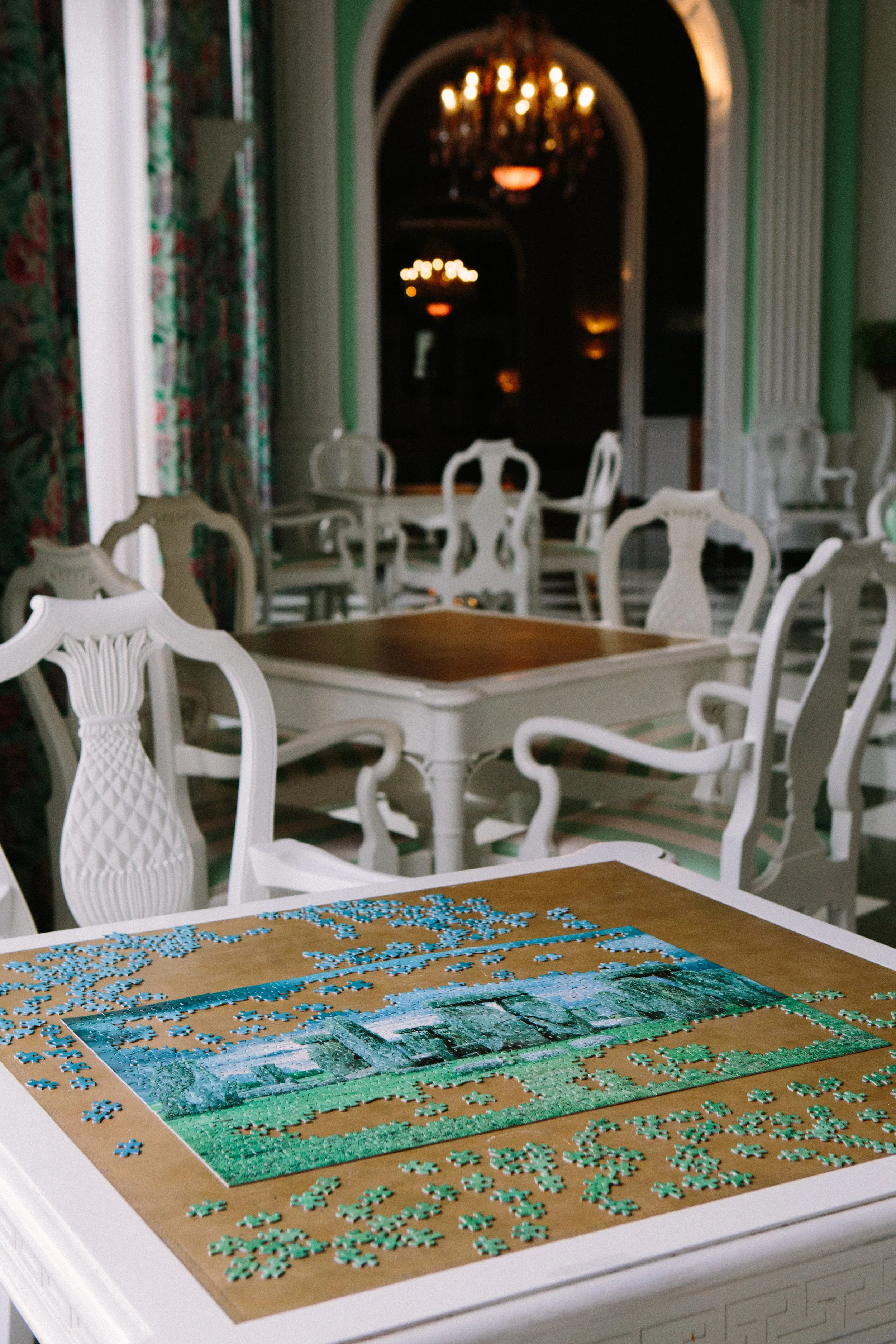 A partially completed jigsaw puzzle of a landscape scene with grass and water is on a table. The puzzle pieces are scattered around it in an indoor setting with elegant white chairs and a large archway with green walls and floral curtains.
