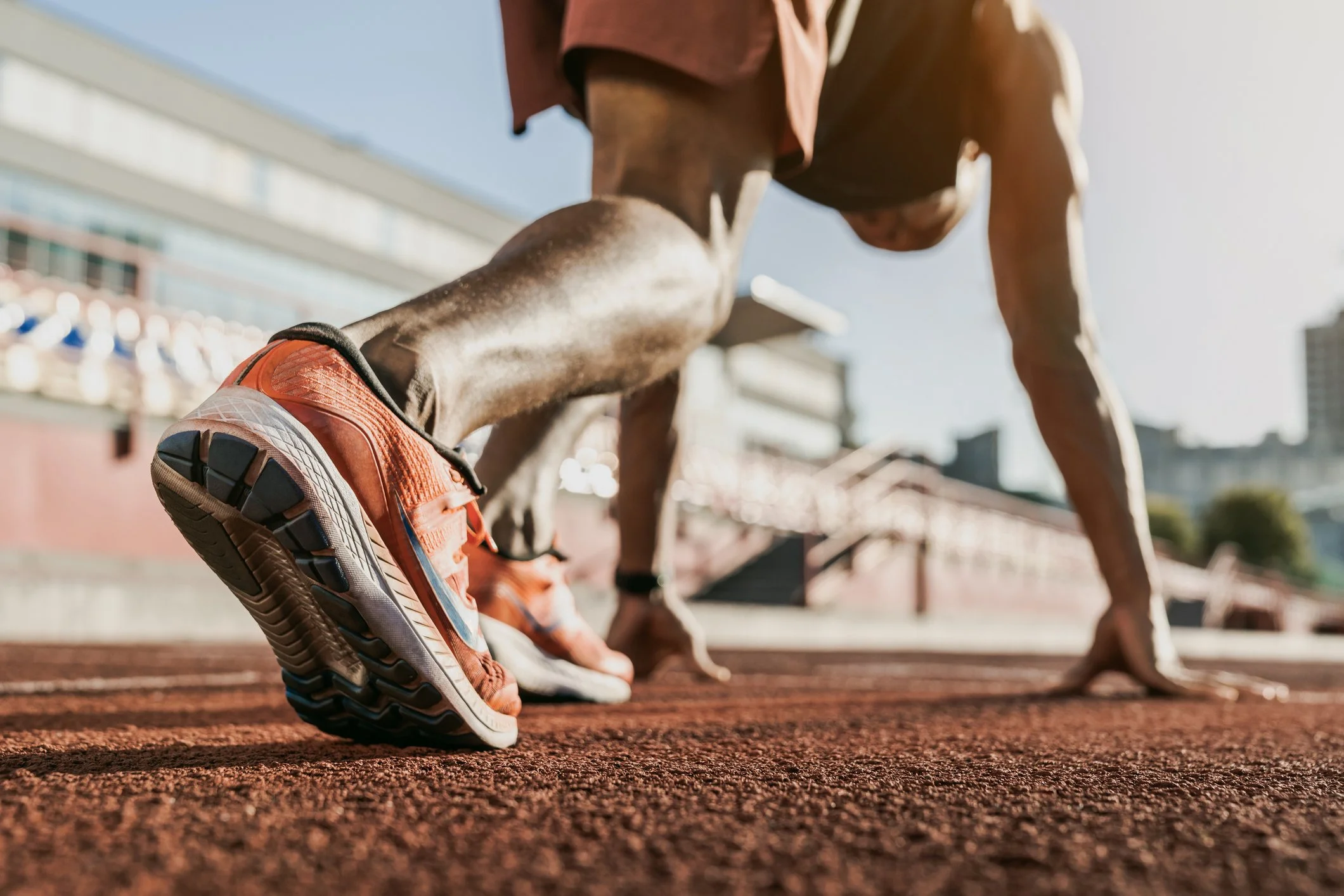 Close-up of a runner's orange athletic shoe on a red track, with a runner in starting position in the background near a stadium.