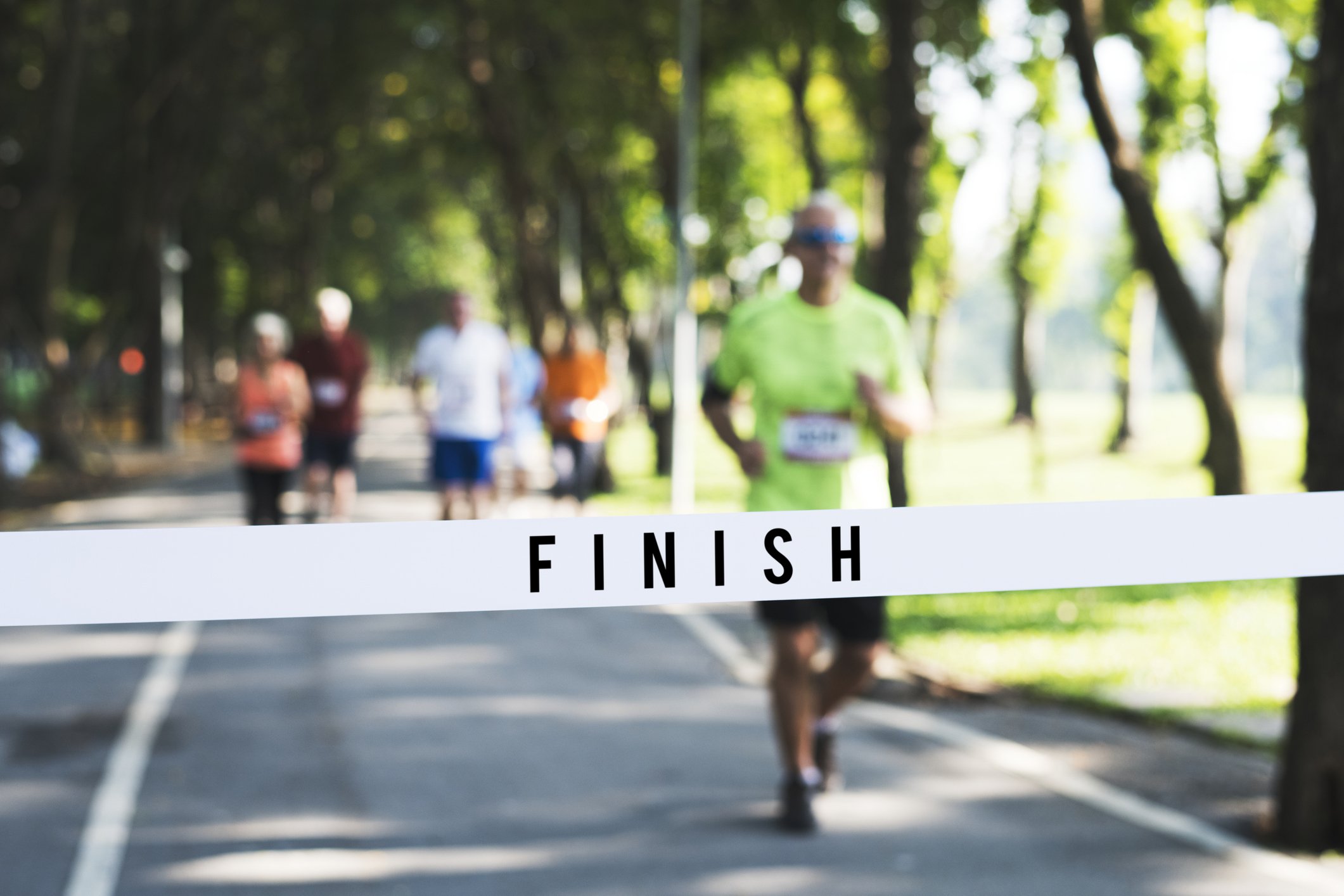 A group of runners crossing the finish line in a race, with a person in a bright green shirt and blue sunglasses leading.