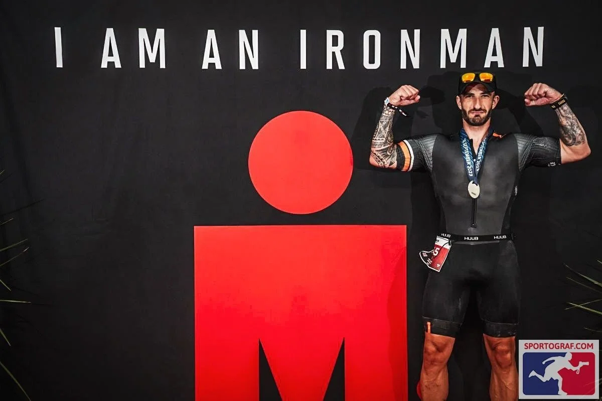 Man with a beard wearing a medal, flexing his arms in front of a black background with large text that reads 'I AM AN IRONMAN' and a red logo. The man has tattoos on his arms and is dressed in athletic gear, including a black cap with yellow sunglasses on top, a black vest, and black shorts.