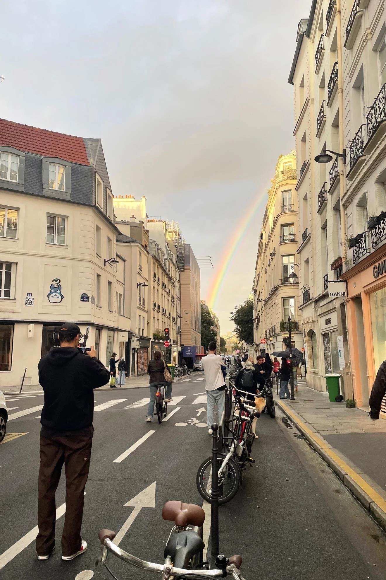 A street scene in a city with buildings on both sides and a rainbow in the sky. People are walking, biking, and taking photos, with parked bicycles and motorcycles along the sidewalk.