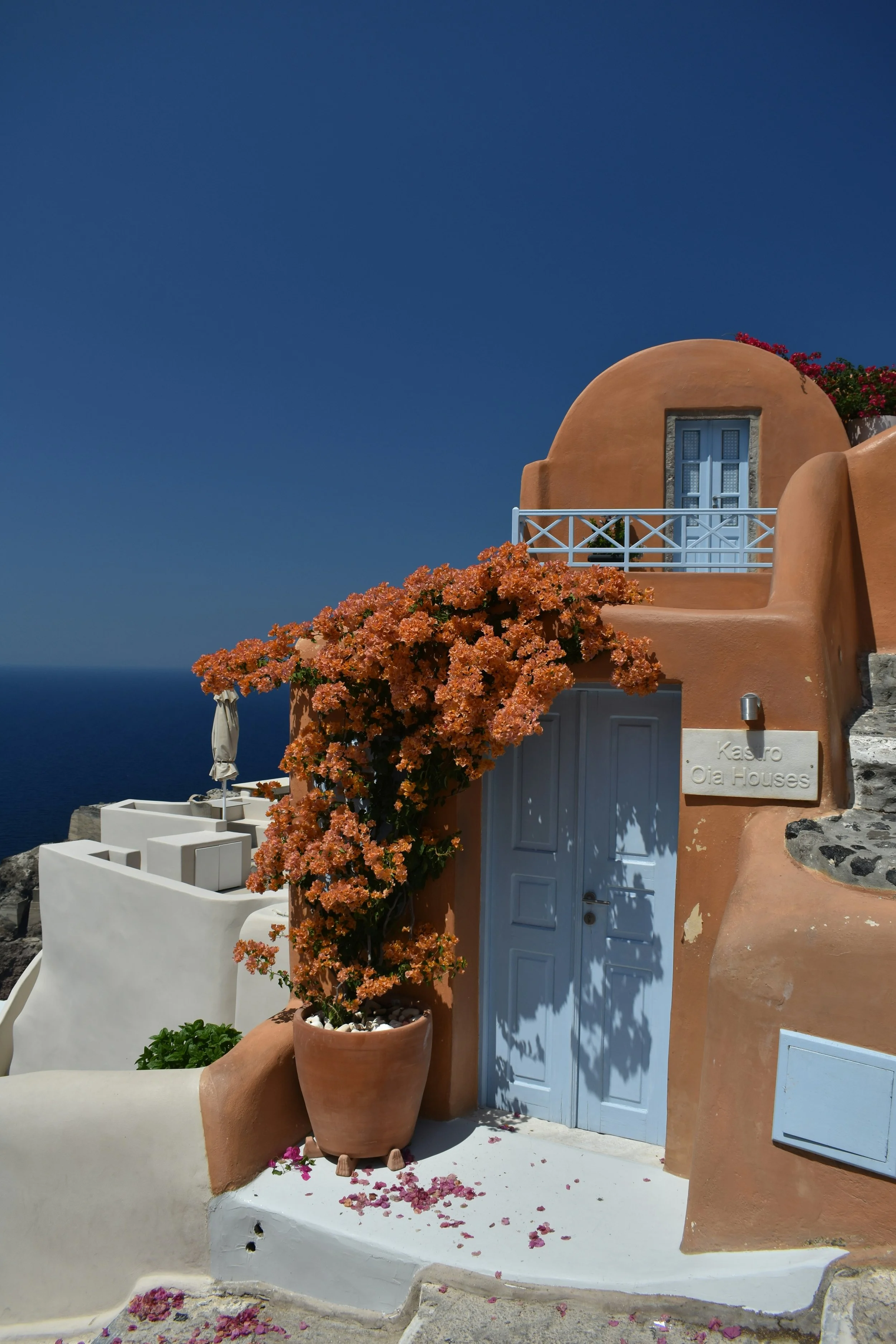 A traditional pinkish-orange building with a curved roof and a blue door, decorated with flowering plants and a white fence, under a clear blue sky.