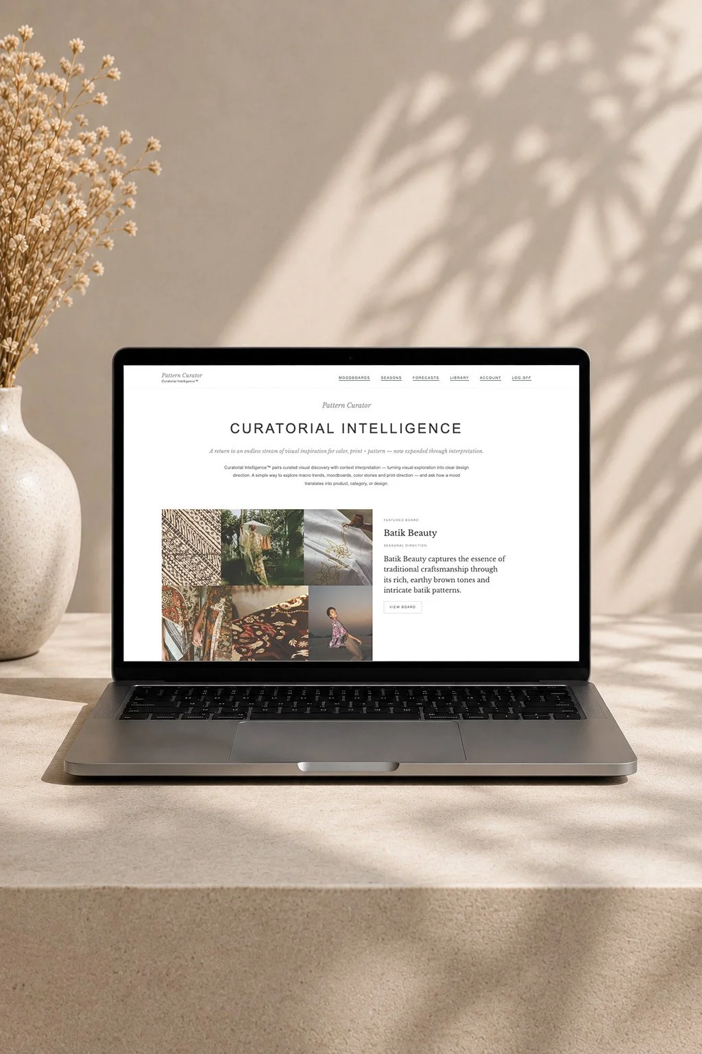 A silver laptop displaying a webpage about curatorial intelligence, placed on a beige table beside a white textured vase with dried flowers, with soft shadows on the background wall.