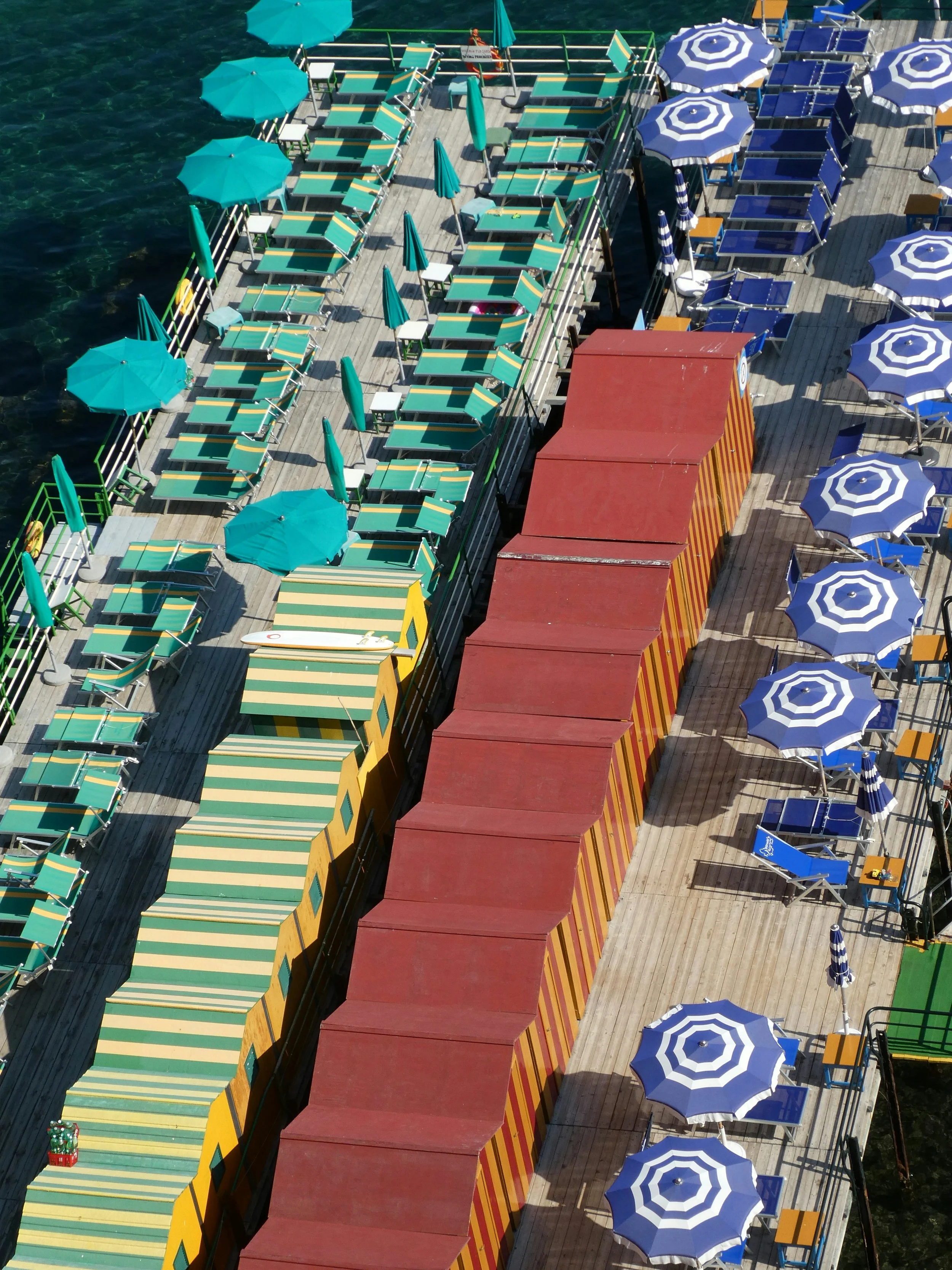 Aerial view of a wooden pier with colorful striped umbrellas, lounge chairs, tables, and a small red and yellow striped structure, over water.