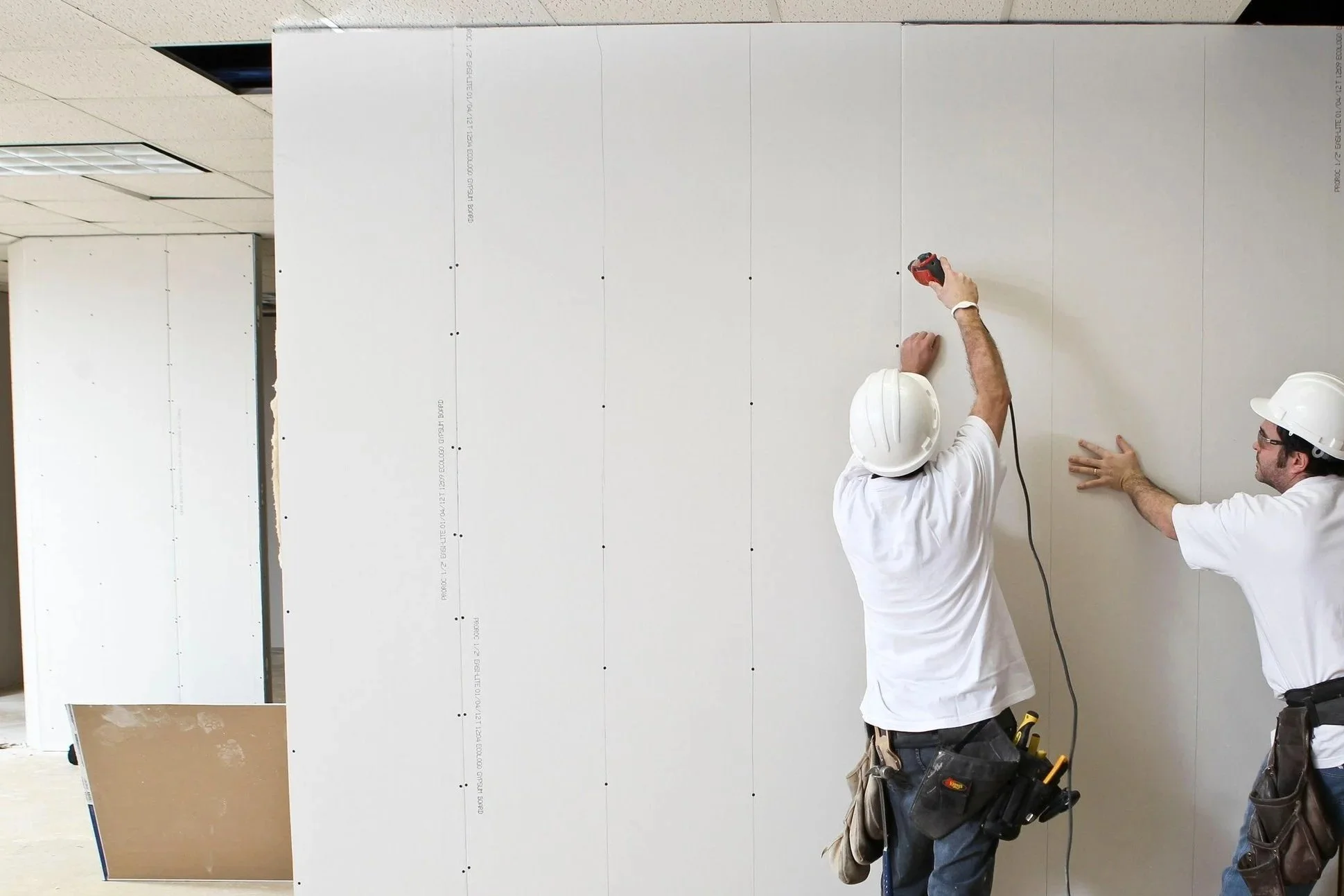 Dos trabajadores con cascos blancos instalando paneles de yeso en una pared interior, uno de ellos sostiene una herramienta eléctrica.