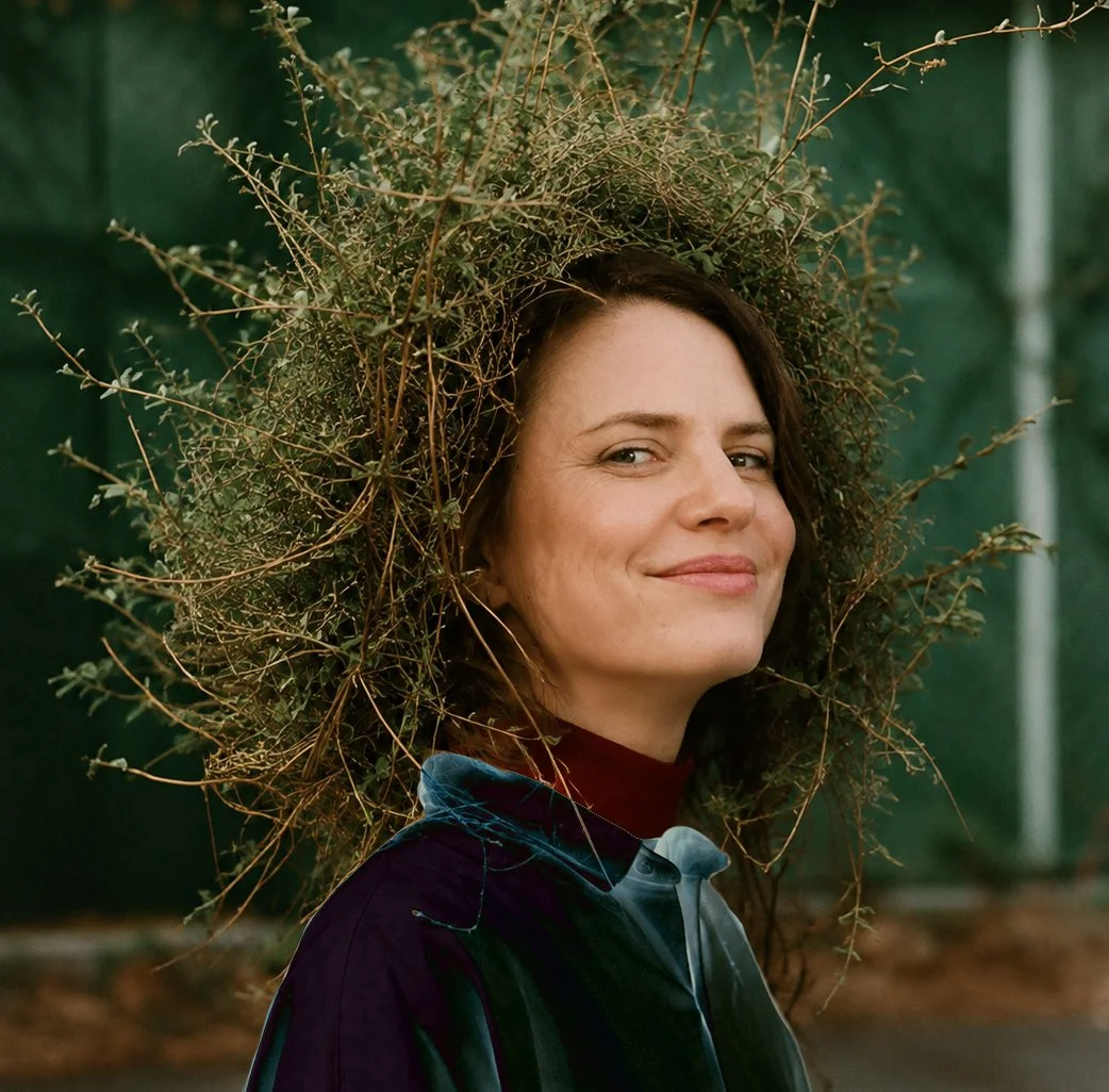 A woman smiling with a large, leafy arrangement around her head, resembling a crown or halo, standing outdoors against a green background.