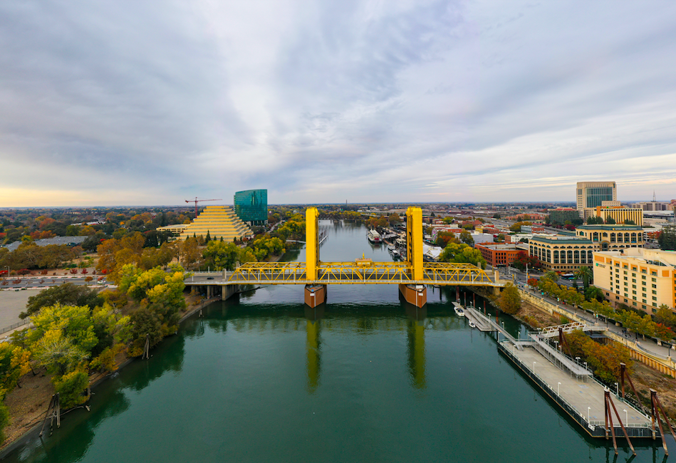 Aerial view of a city with a yellow drawbridge over a river, surrounded by buildings and trees with autumn foliage under a cloudy sky.