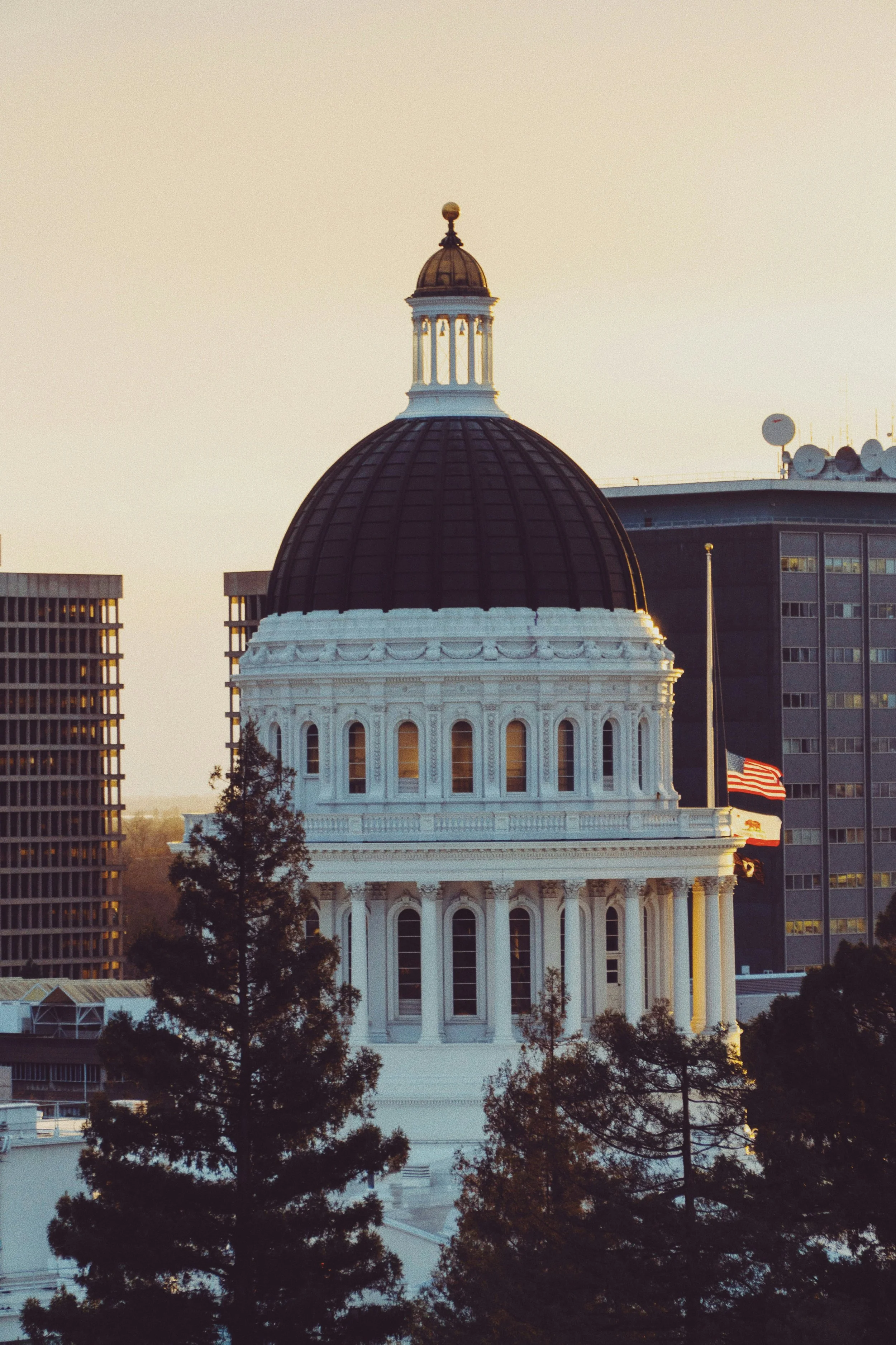 The California State Capitol building in Sacramento, California during sunset, with trees in the foreground and adjacent office buildings, American flag, and TV antennas in the background.