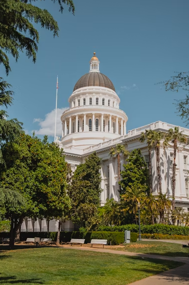 The California State Capitol building in Sacramento, California, with a large dome, surrounded by trees and a landscaped park.