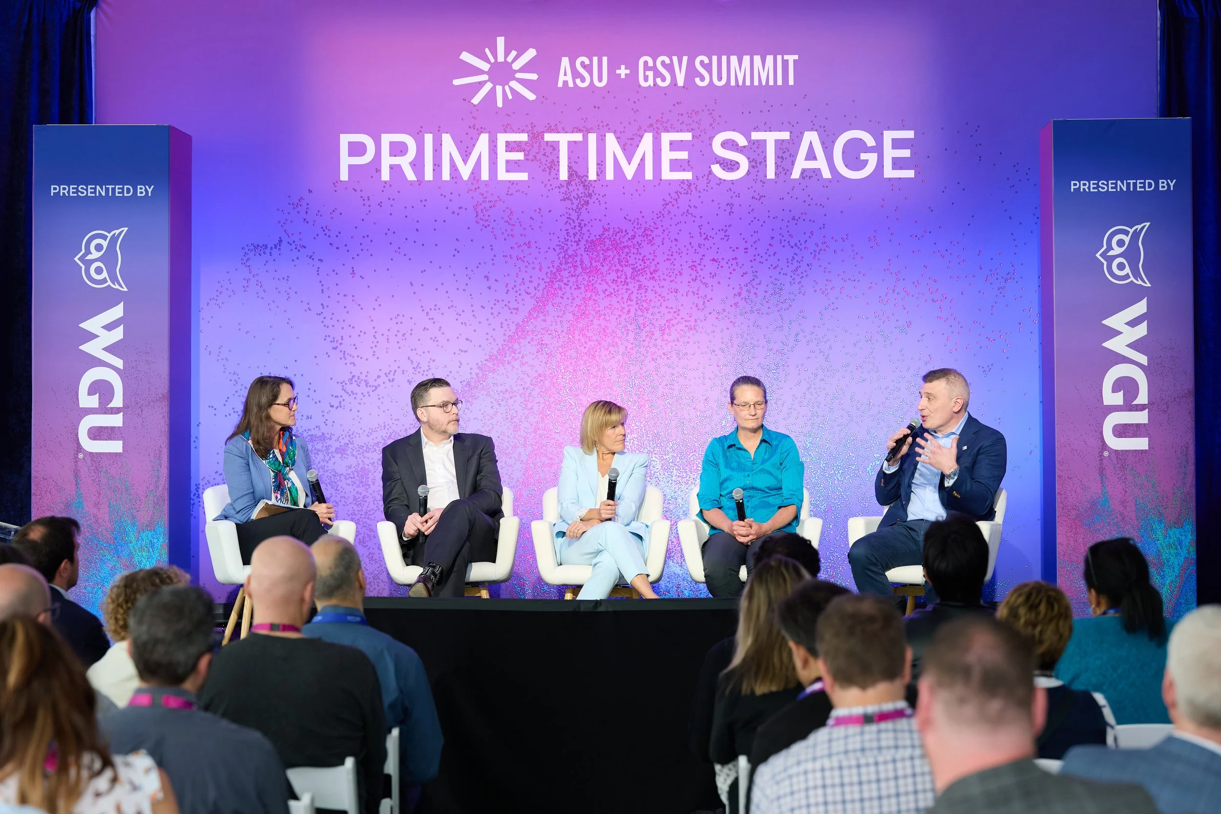 A panel discussion at the ASU + GSV Summit on a stage with Tommy Bishop into a microphone, with a purple backdrop displaying ASU+GSV Logo.