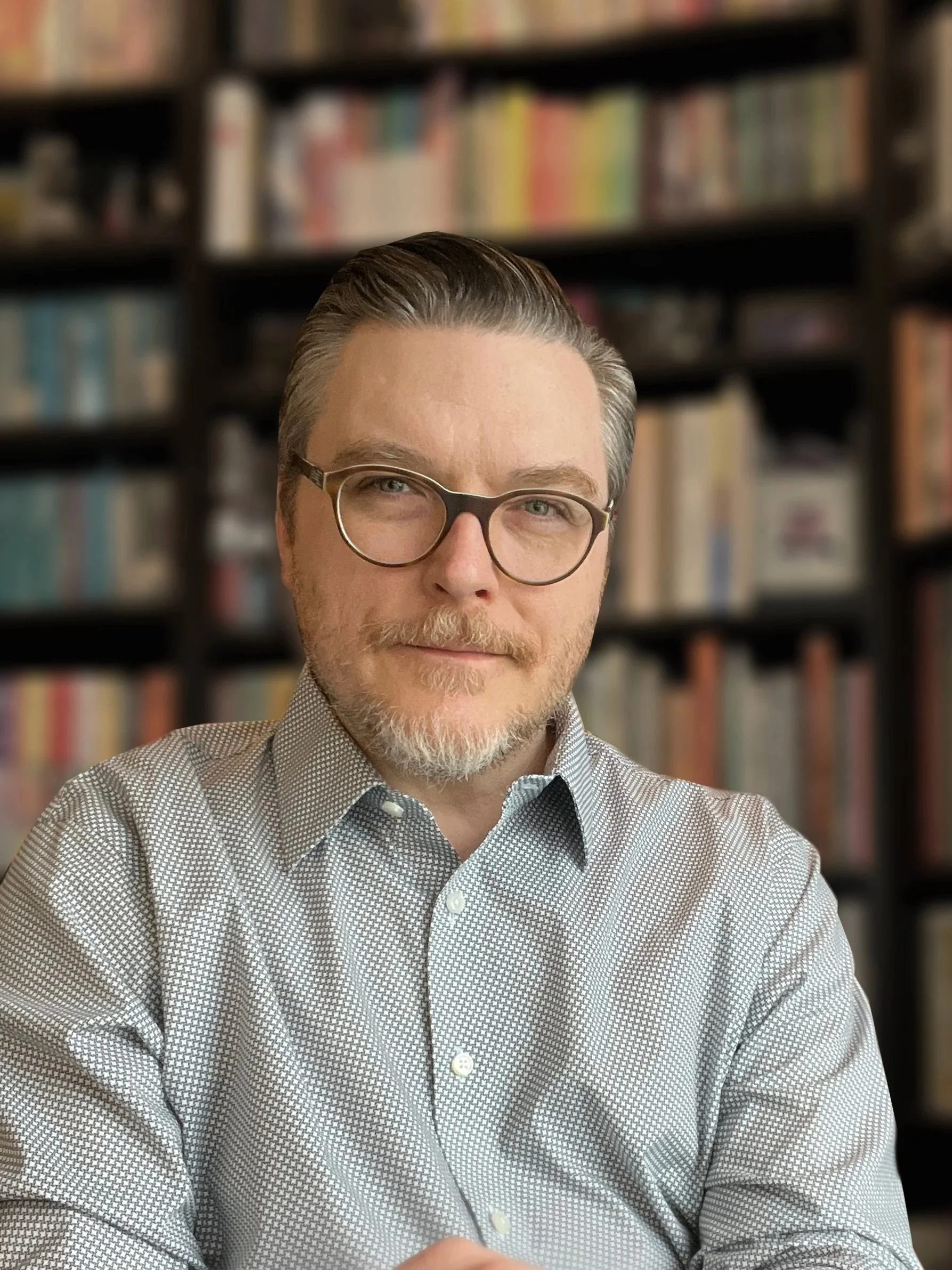 A man with glasses and a beard wearing a gray patterned shirt, sitting in front of a background of bookshelves filled with colorful books.