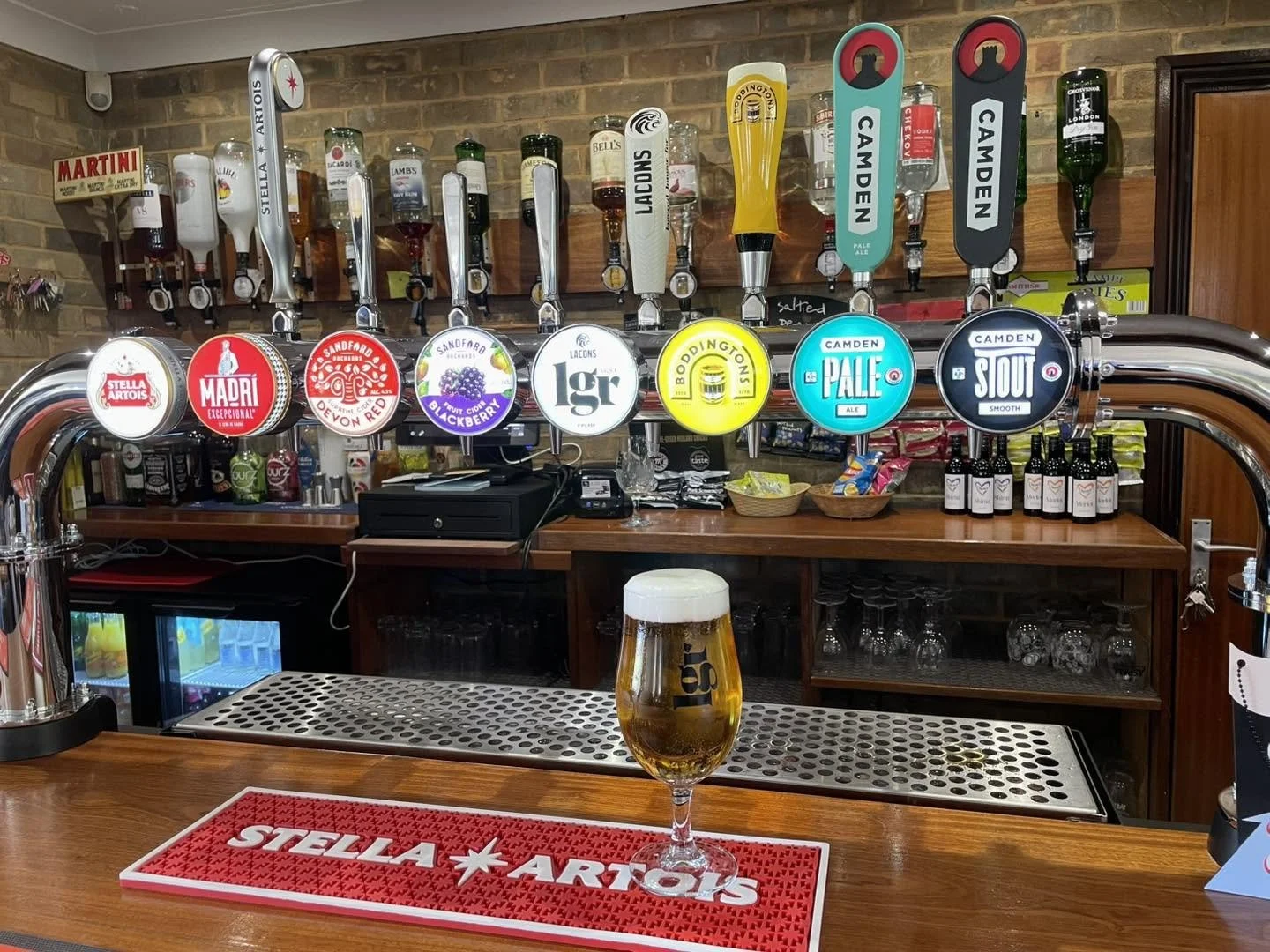 A bar with eight beer taps, each labeled with different beer brands and styles, behind a wooden counter with a glass of beer in the foreground. The bar has a brick wall background with beverage bottles and snacks.