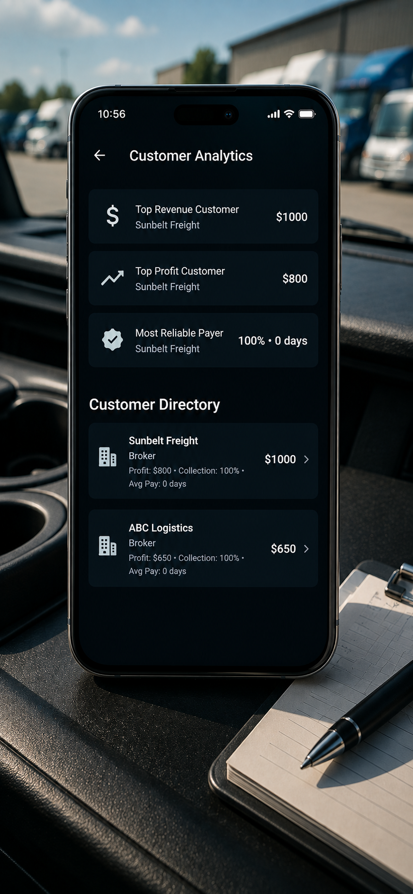 Smartphone displaying customer analytics and customer directory for freight companies, placed on a desk inside a vehicle with a notepad and pen nearby.