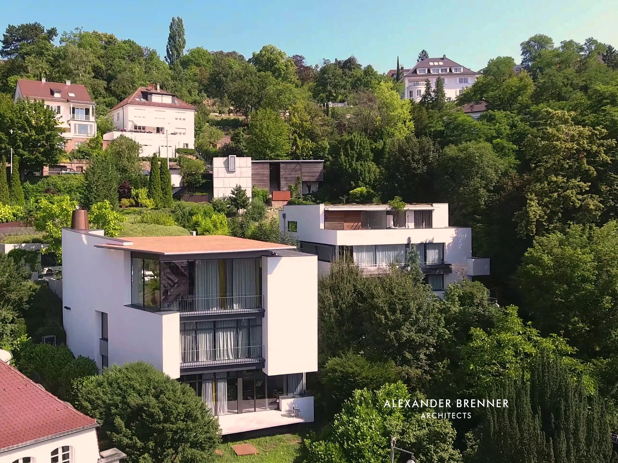 Moderne weiße Villa mit großen Fenstern und Balkon im Grünen, umgeben von Bäumen und anderen Häusern, bei sonnigem Himmel in einer beherrschten Nachbarschaft.