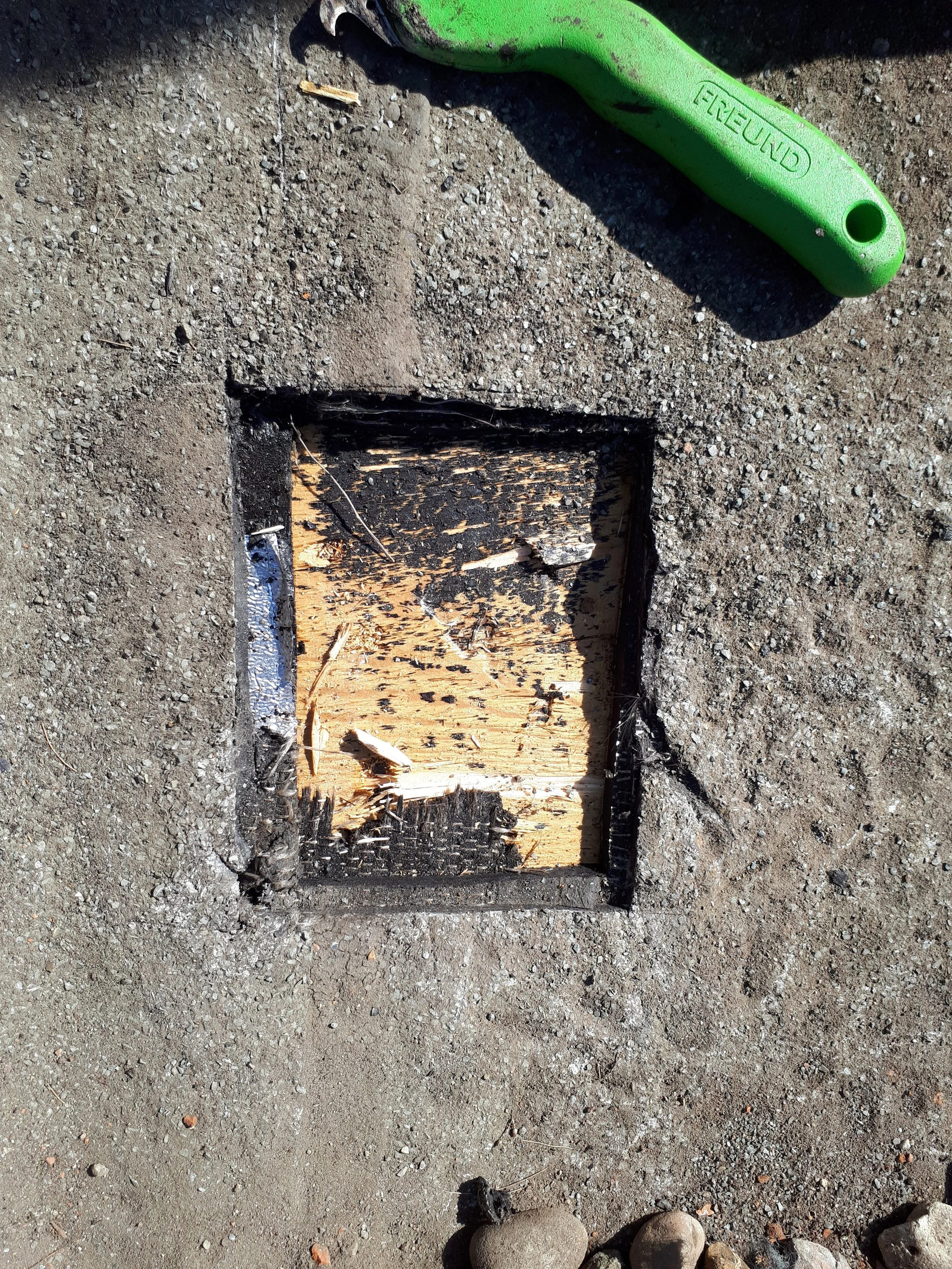Das Bild zeigt ein geöffnetes Loch im Boden, das die Holzstruktur eines Fundaments zeigt. Daneben liegt ein grüner Schraubenschlüssel auf dem Boden.