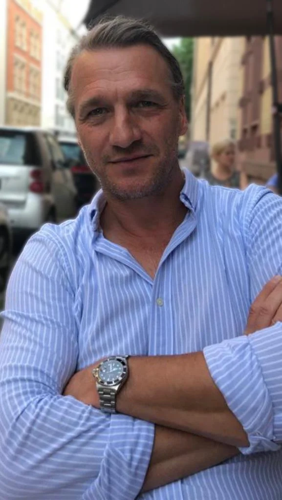 Ein Mann mit kurz braunem Haar, blauer gestreifter Hemd, trägt eine Armbanduhr und steht vor einer belebten Stadtstraße mit Autos und Gebäuden.