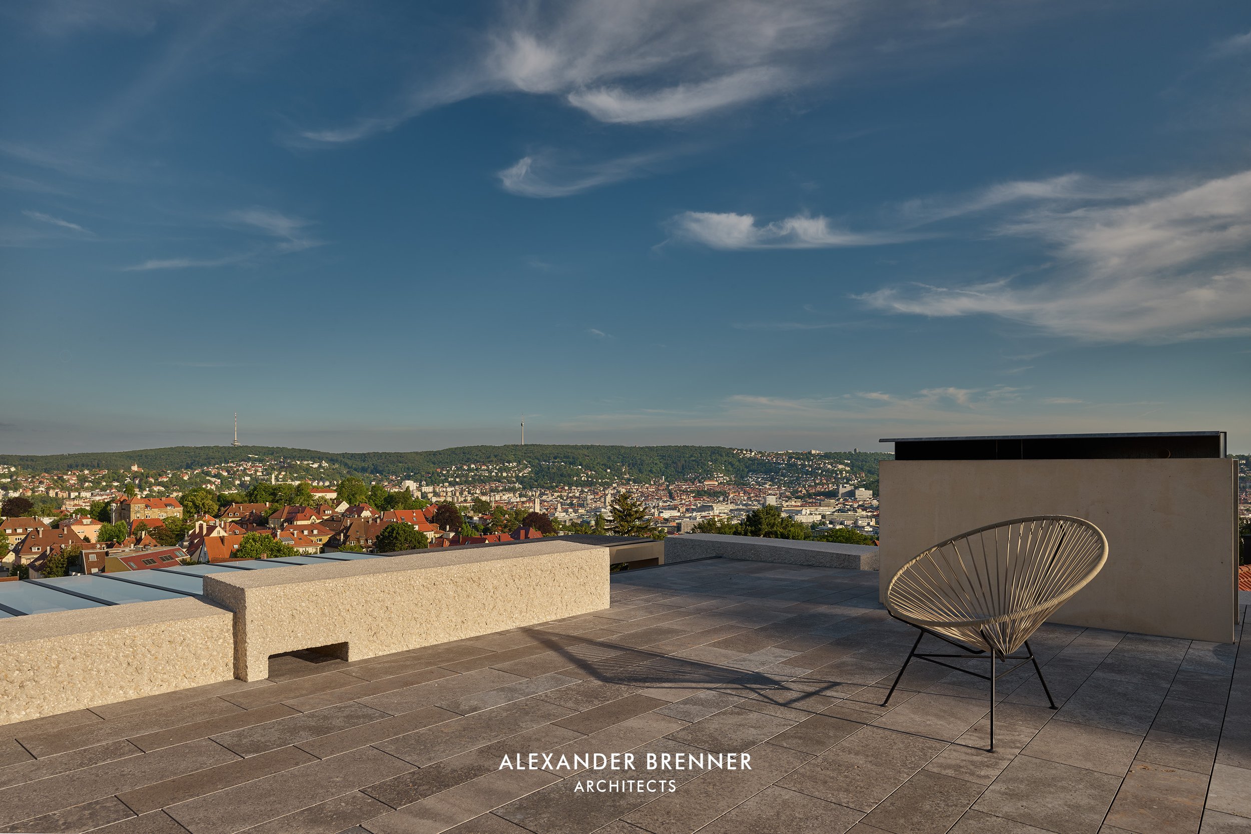 Blick von einer Dachterrasse über eine Stadt mit Häusern, Bäumen und Hügeln im Hintergrund. Das Bild zeigt den Himmel mit einigen Wolken.