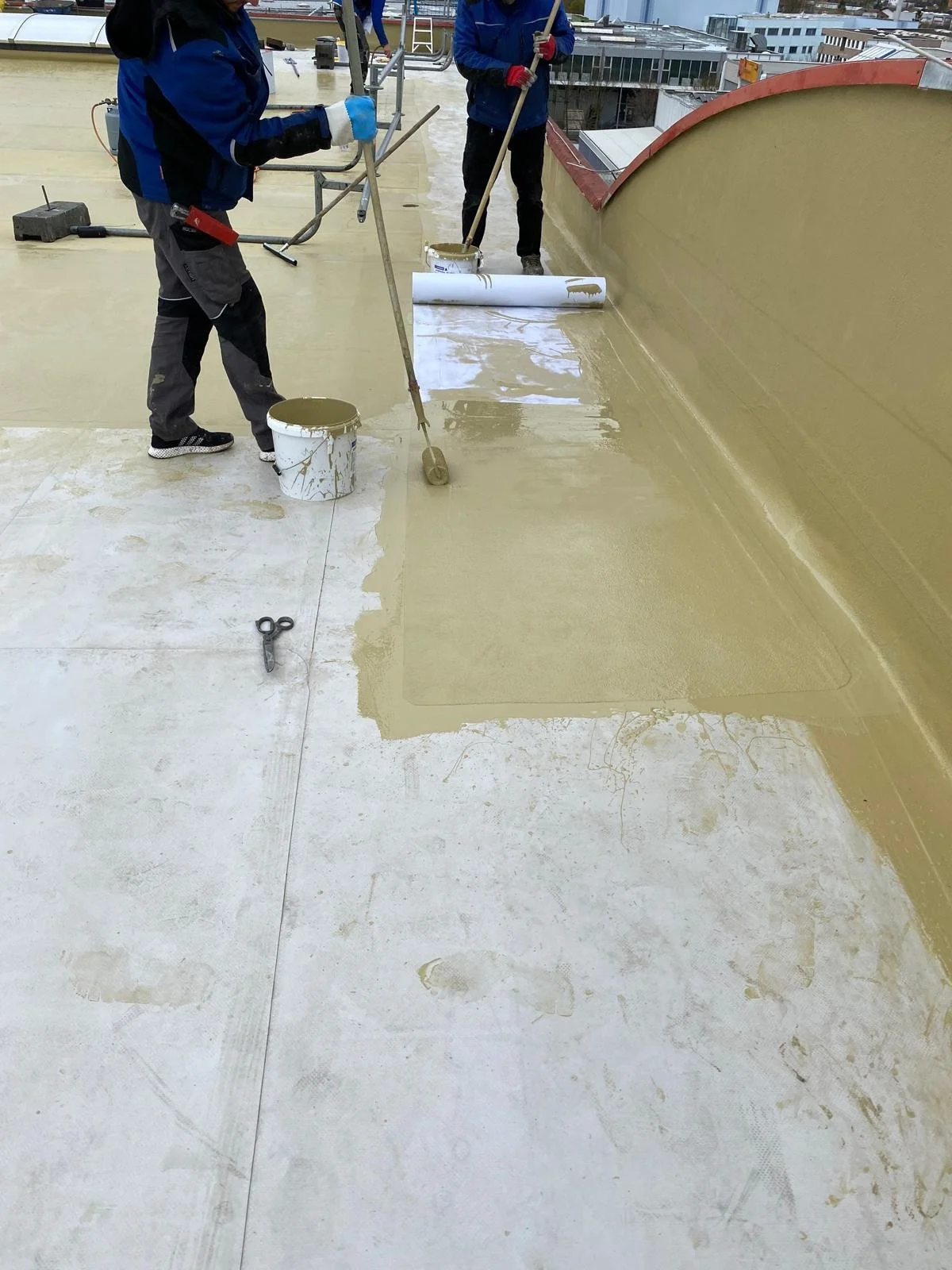 Arbeiter streichen eine Dachterrasse mit Flüßigkunststoff, Werkzeuge und Eimer sind sichtbar.