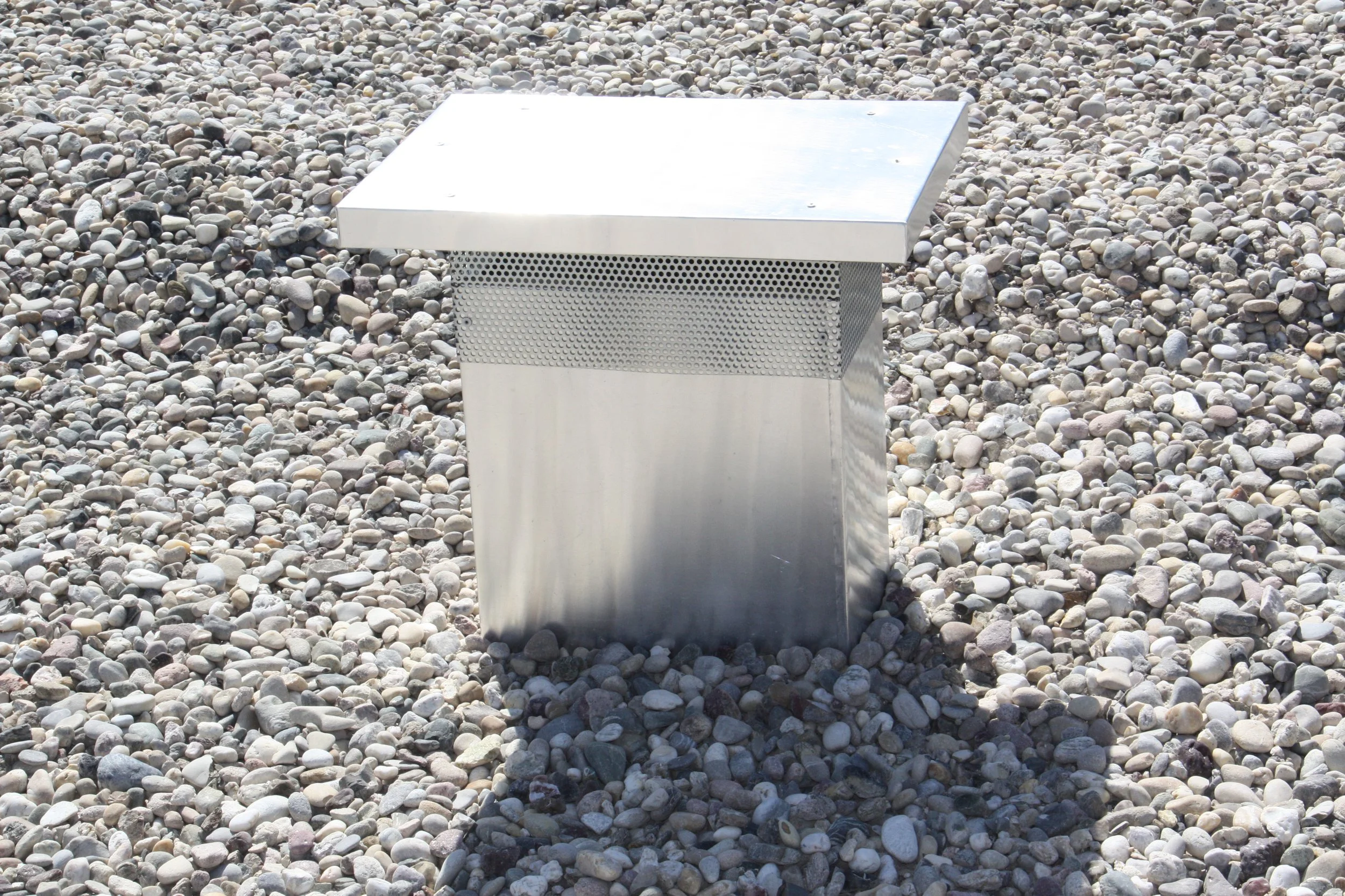 Eine metallene, quadratische Struktur auf einer Steinterrasse, umgeben von Kieselsteinen.