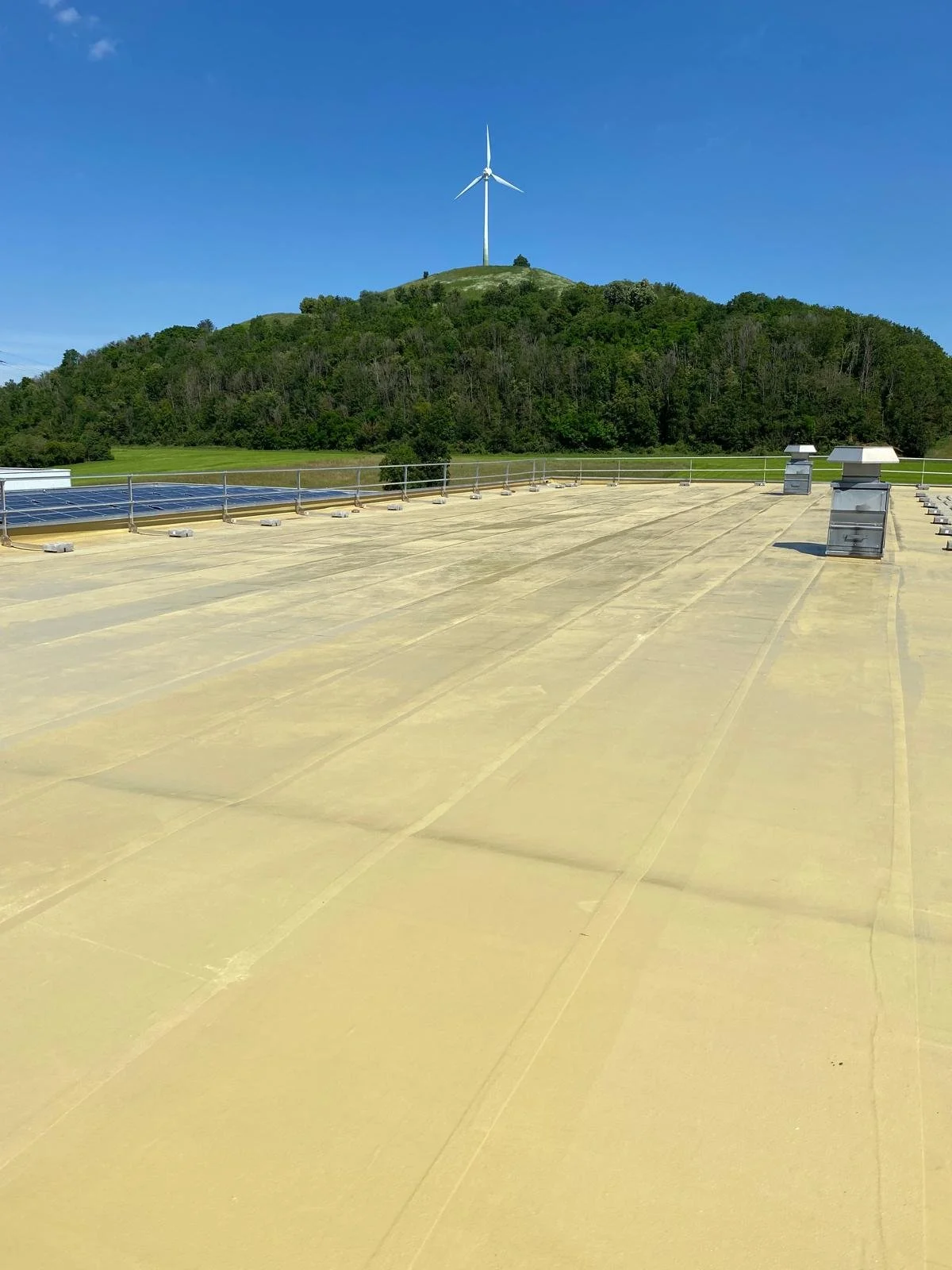 Auf einer flachen Dachfläche sind mehrere Dachventilatoren und eine Solaranlage zu sehen. Im Hintergrund befindet sich ein grüner Hügel mit einem Windrad auf der Spitze, umgeben von Bäumen und bewölktem Himmel.