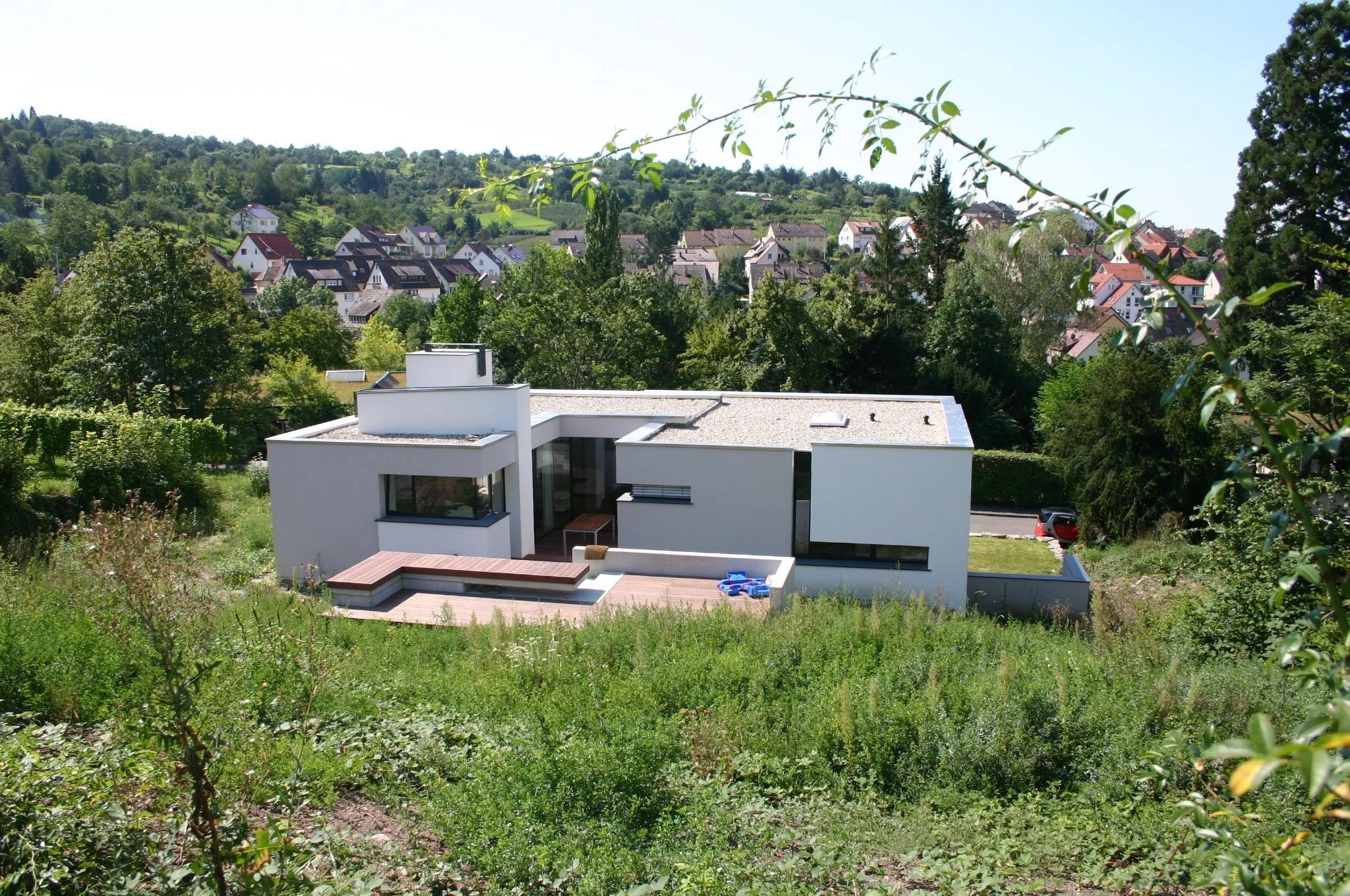 Moderne weiße Villa mit Terrasse, umgeben von grünem Garten und Bäumen, mit Blick auf ein Dorf in einer hügeligen Landschaft.