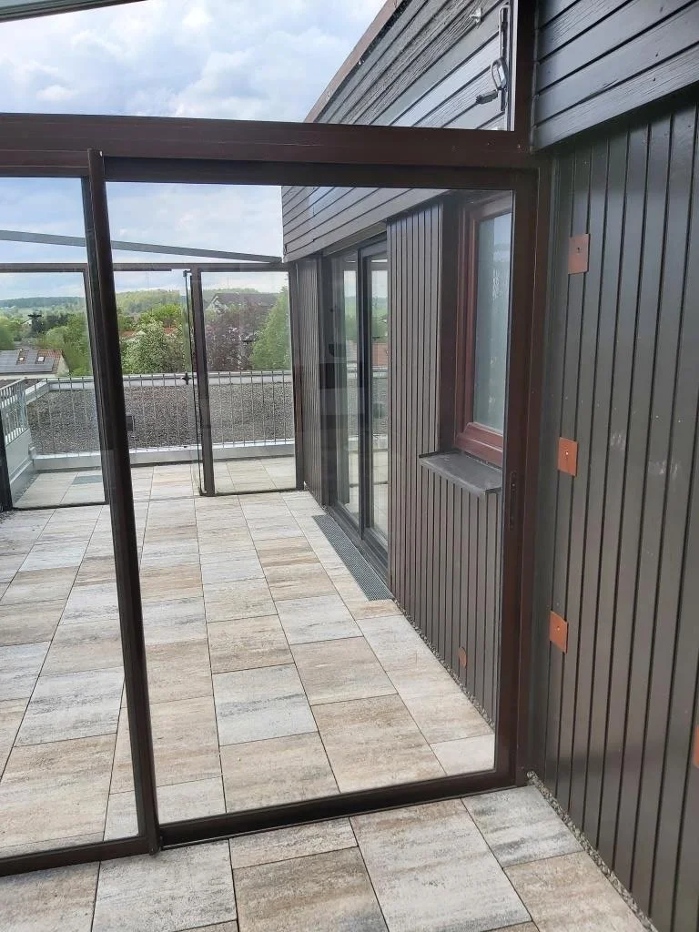 Blick von einem Balkon mit Glasgeländer auf eine grüne Landschaft mit Bäumen und Häusern im Hintergrund, bei bewölktem Himmel.