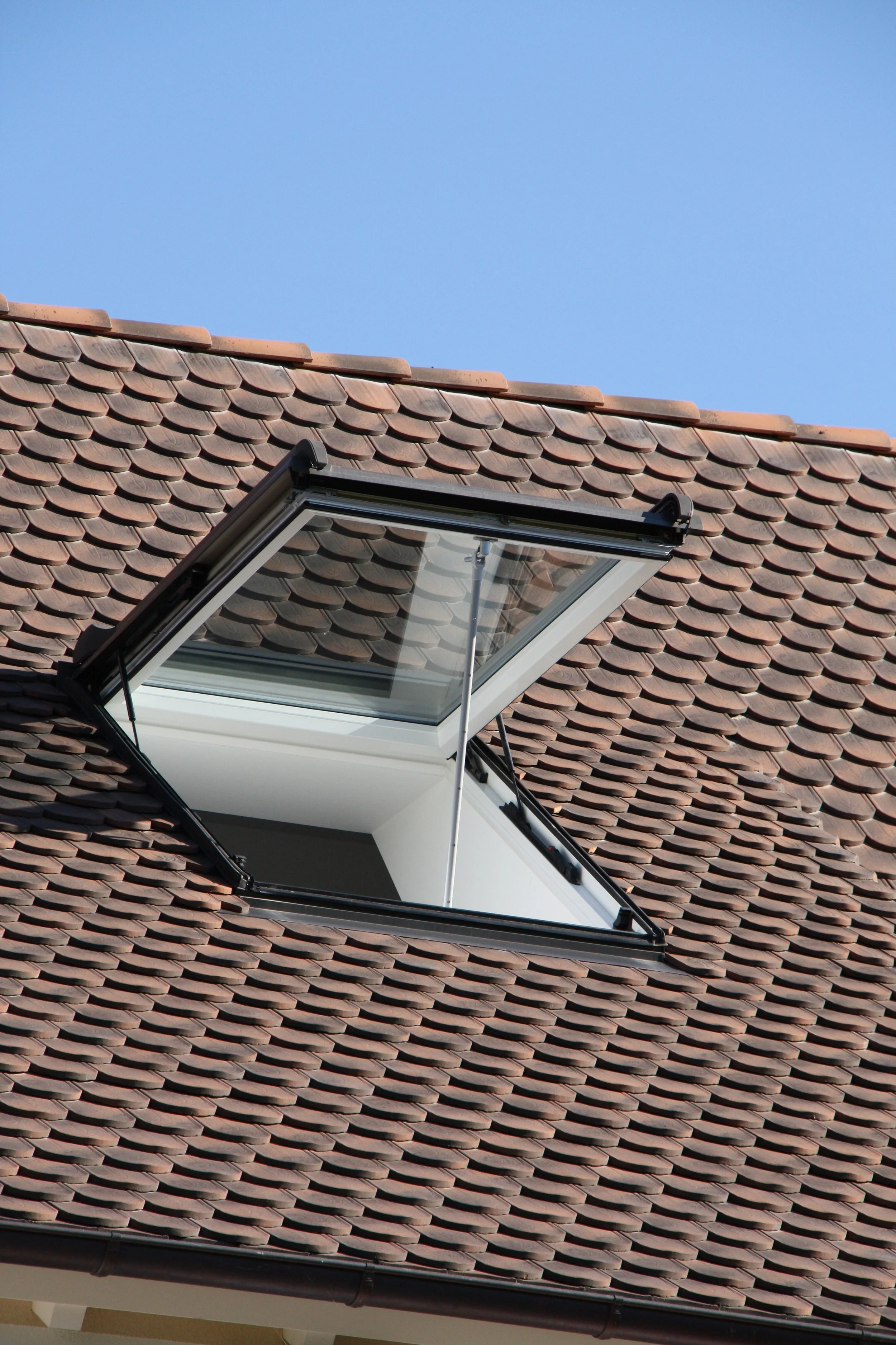 Dach mit roten Ziegeln und offenes Dachfenster, blauer Himmel im Hintergrund.