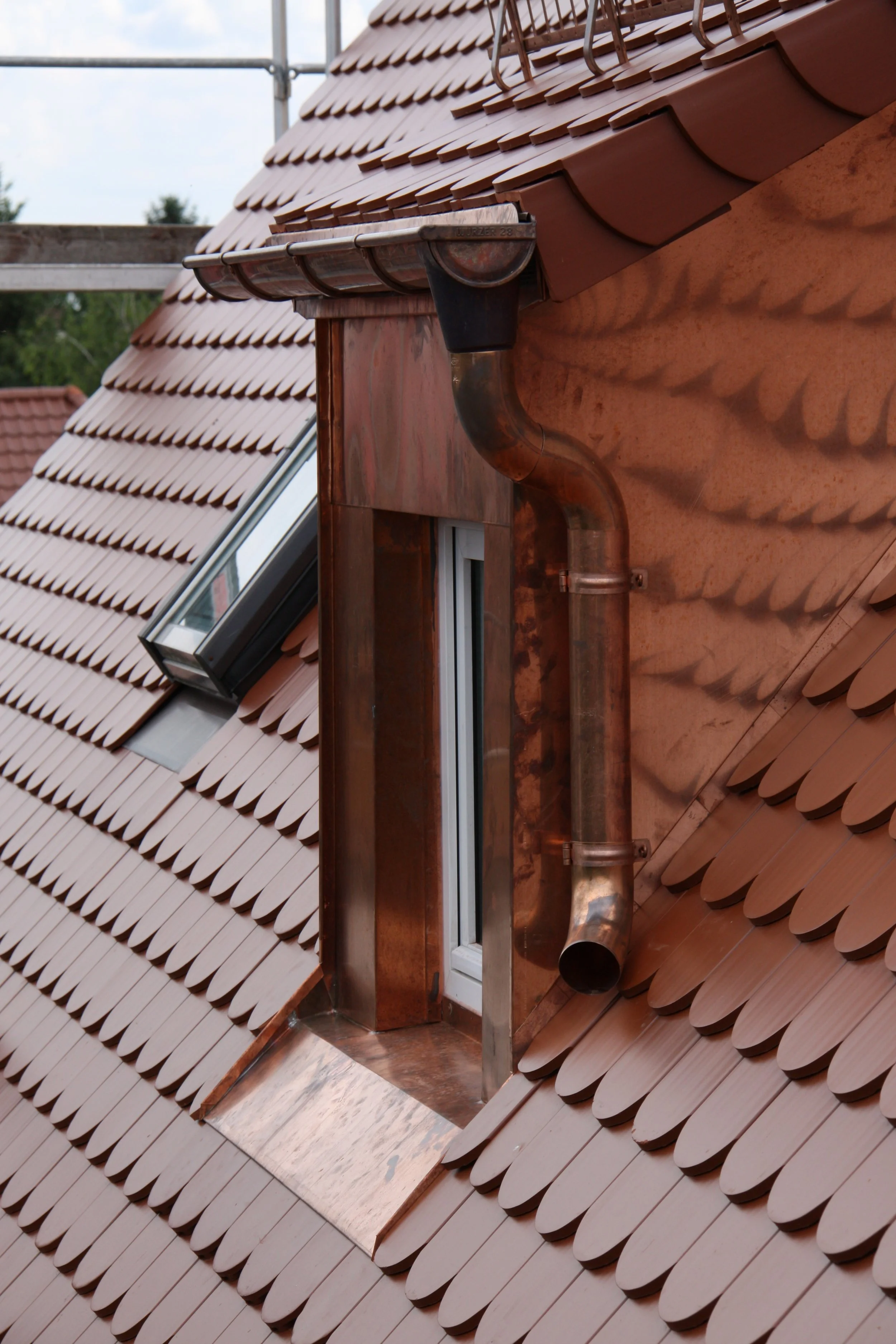 A copper-colored roof with overlapping tiles, a skylight window, and a copper gutter pipe.