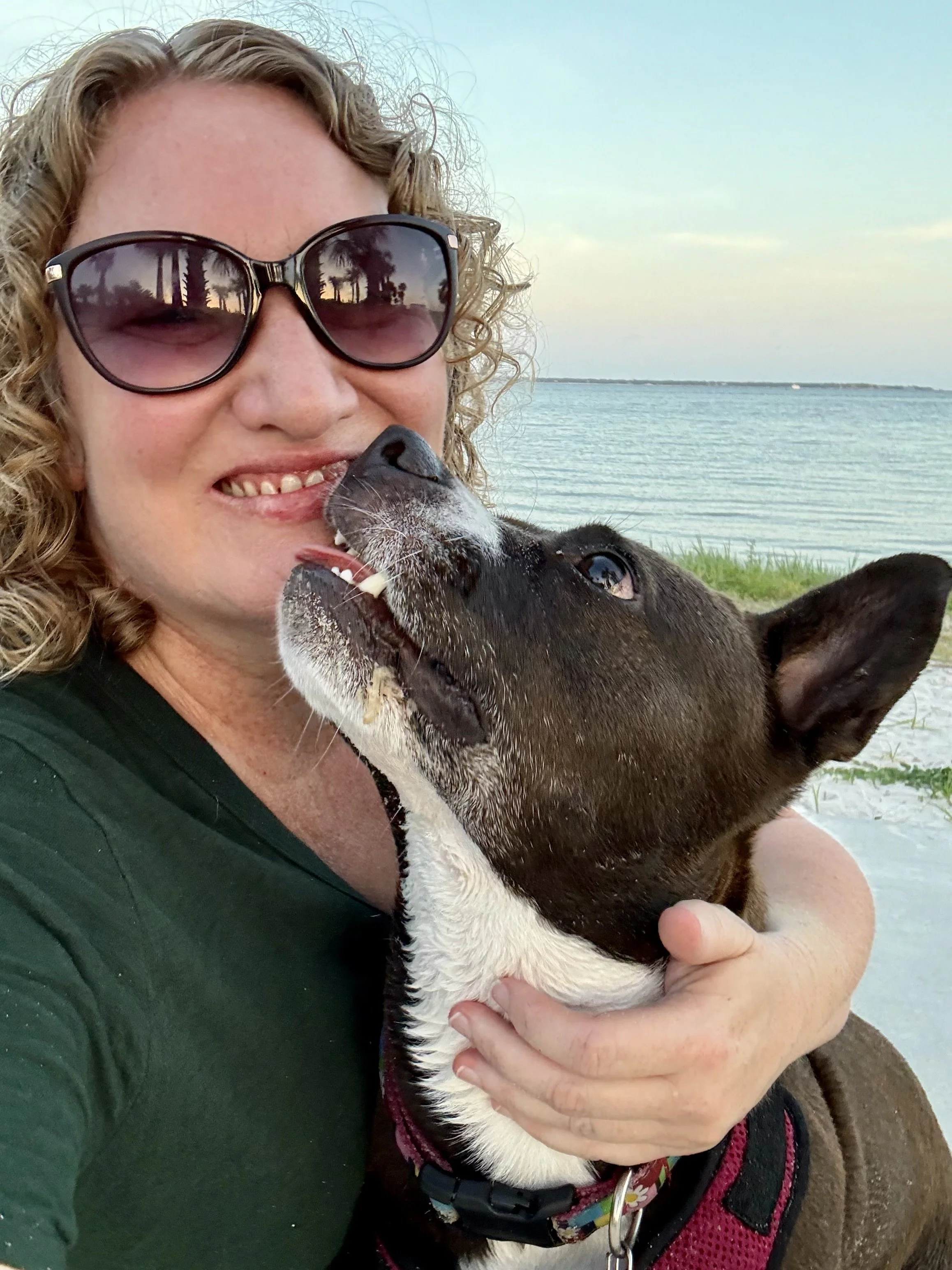 A woman with curly hair and sunglasses holding a black and white dog near a body of water, with a cloudy sky in the background.