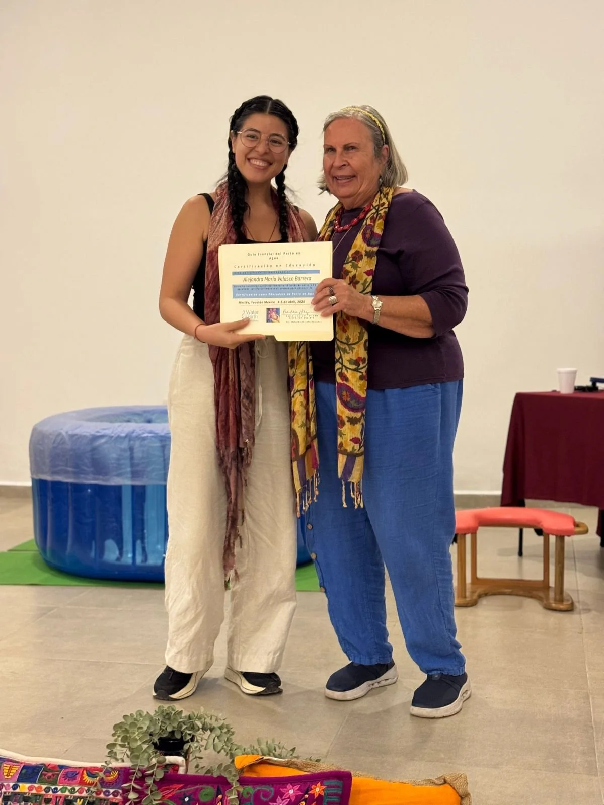 Certified doula Ale proudly holding her training certificate next to an instructor, standing in front of a blue water birth pool.