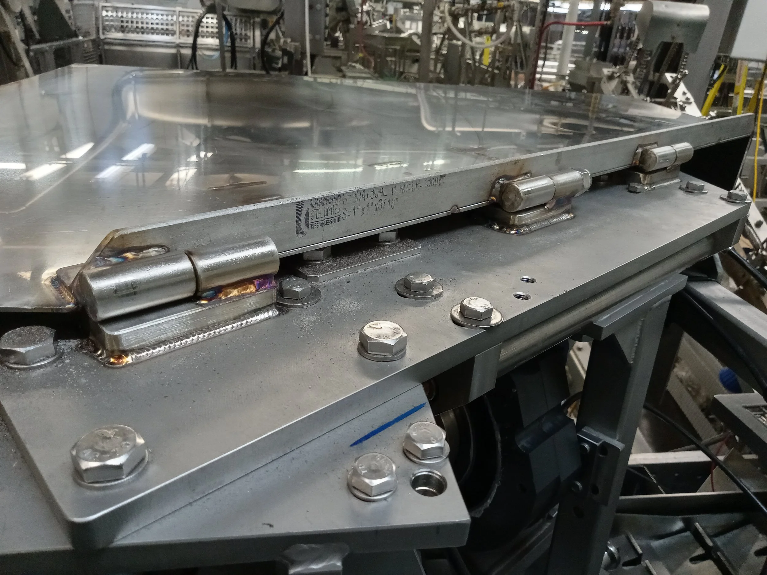 Close-up of a stainless steel industrial mold or metal tray with hydraulic hinges, bolts, and machinery in the background, likely in a manufacturing or industrial setting.