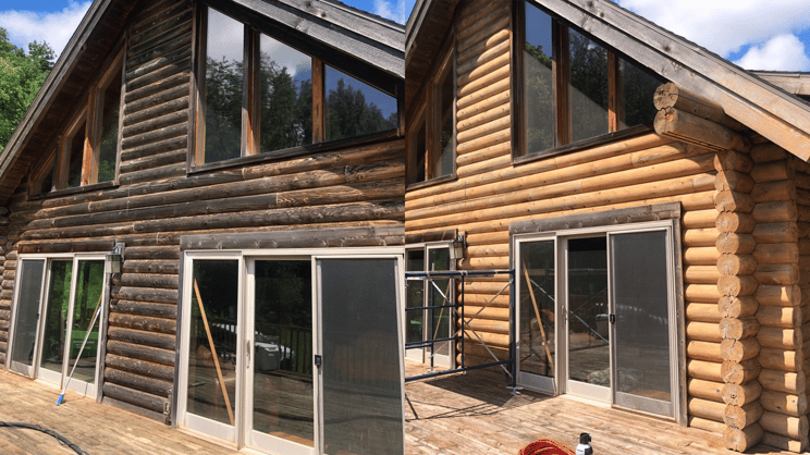 Side-by-side comparison of a log cabin exterior during construction, showing before and after finishes. The left side features dark stained logs with gray sliding glass doors, while the right side has light natural logs with similar sliding doors. Scaffolding and construction equipment are visible, with trees and a clear blue sky in the background.