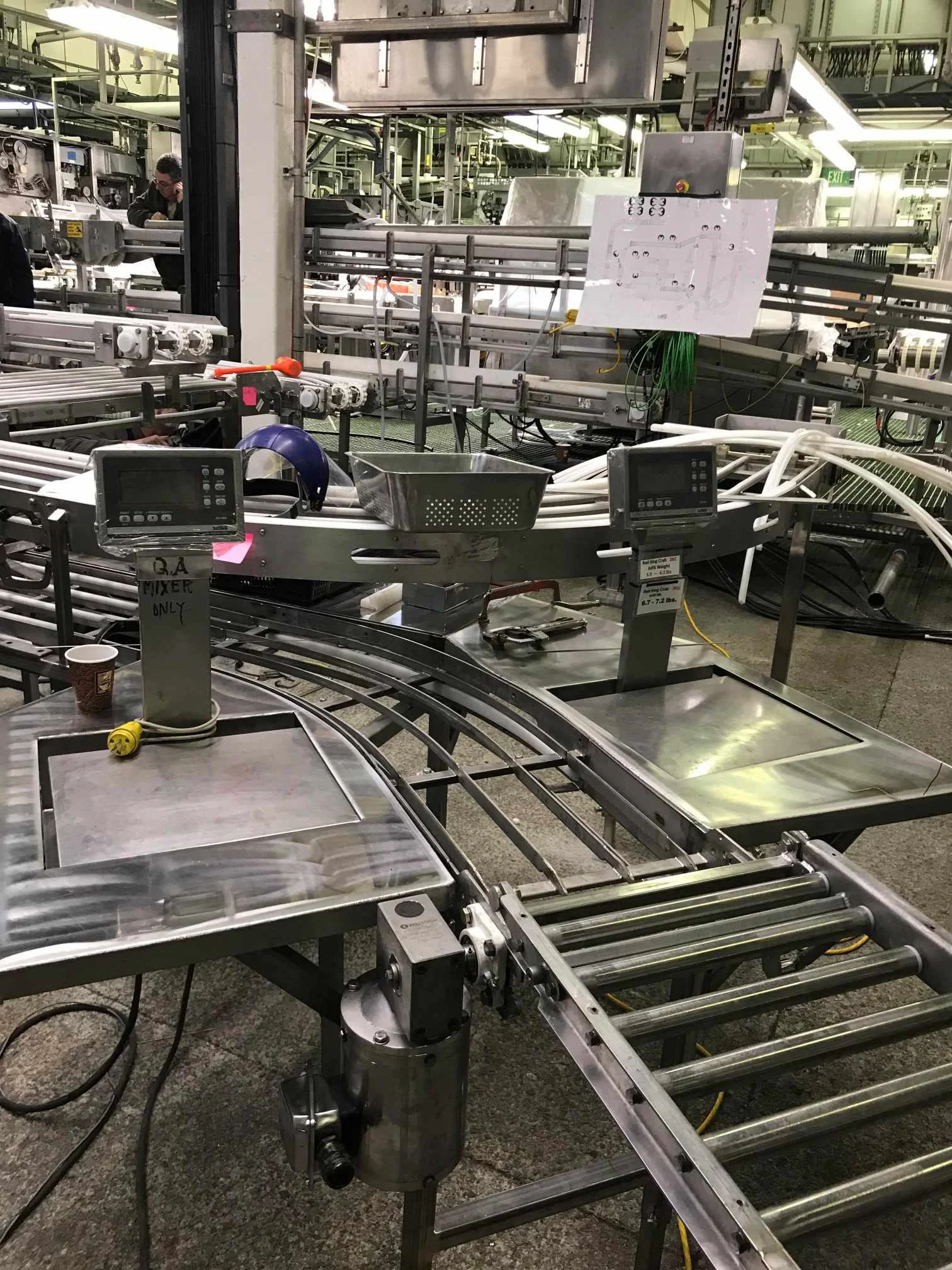 Industrial conveyor belt system with metal rollers, control panels, and wiring, used in manufacturing or packaging environment.
