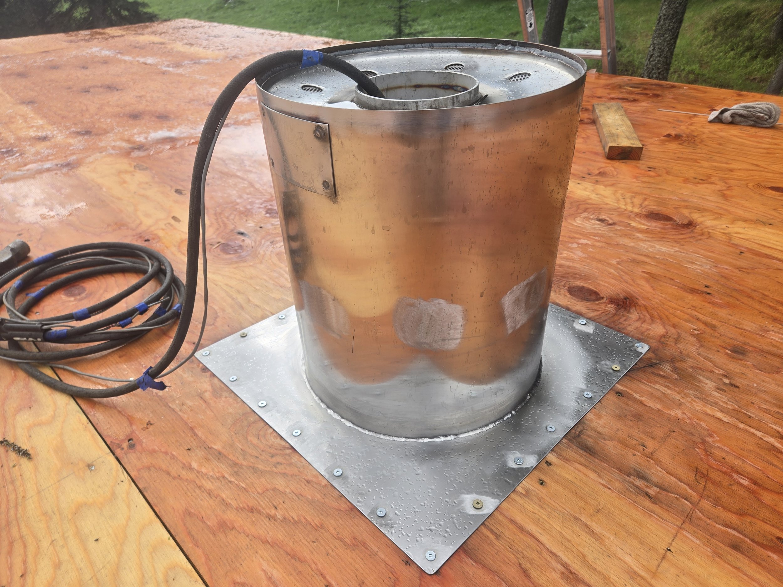 A metallic cylindrical device with a black hose attached, placed on a wooden surface outdoors, with some tools and wires nearby.