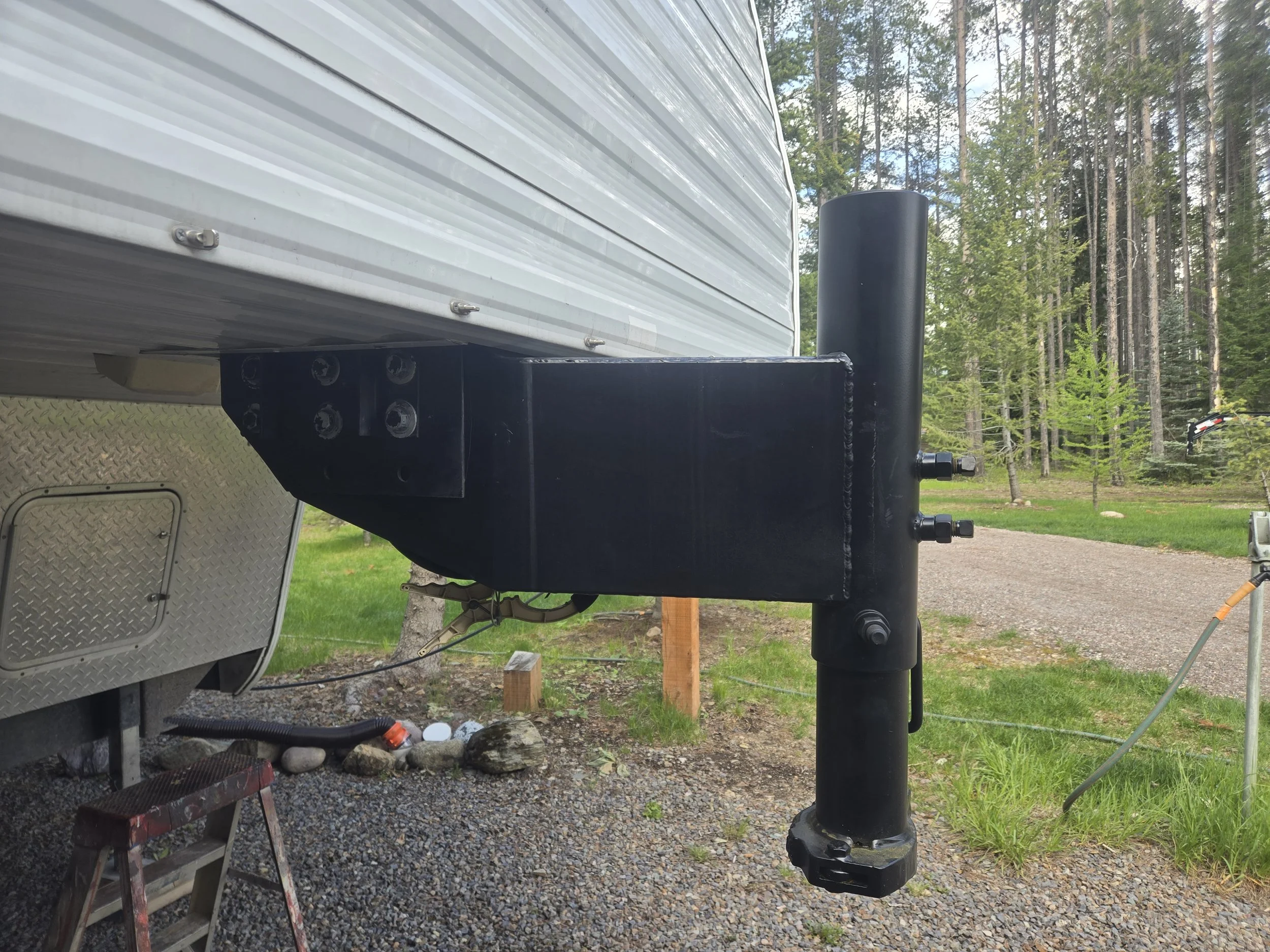 Close-up of a black trailer hitch mounted on a raised platform in a wooded outdoor area, with trees and a trail in the background.