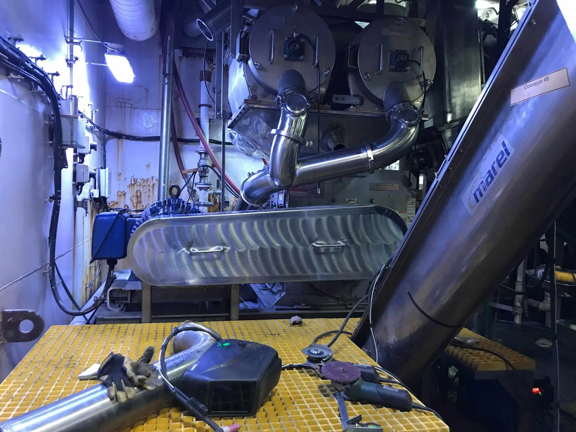 Inside a cleanroom or laboratory with stainless steel equipment, pipes, and tools on a yellow grated surface. Equipment labeled "Conveyor #8" and a cylindrical device are present. The environment appears to be for industrial or technological work.