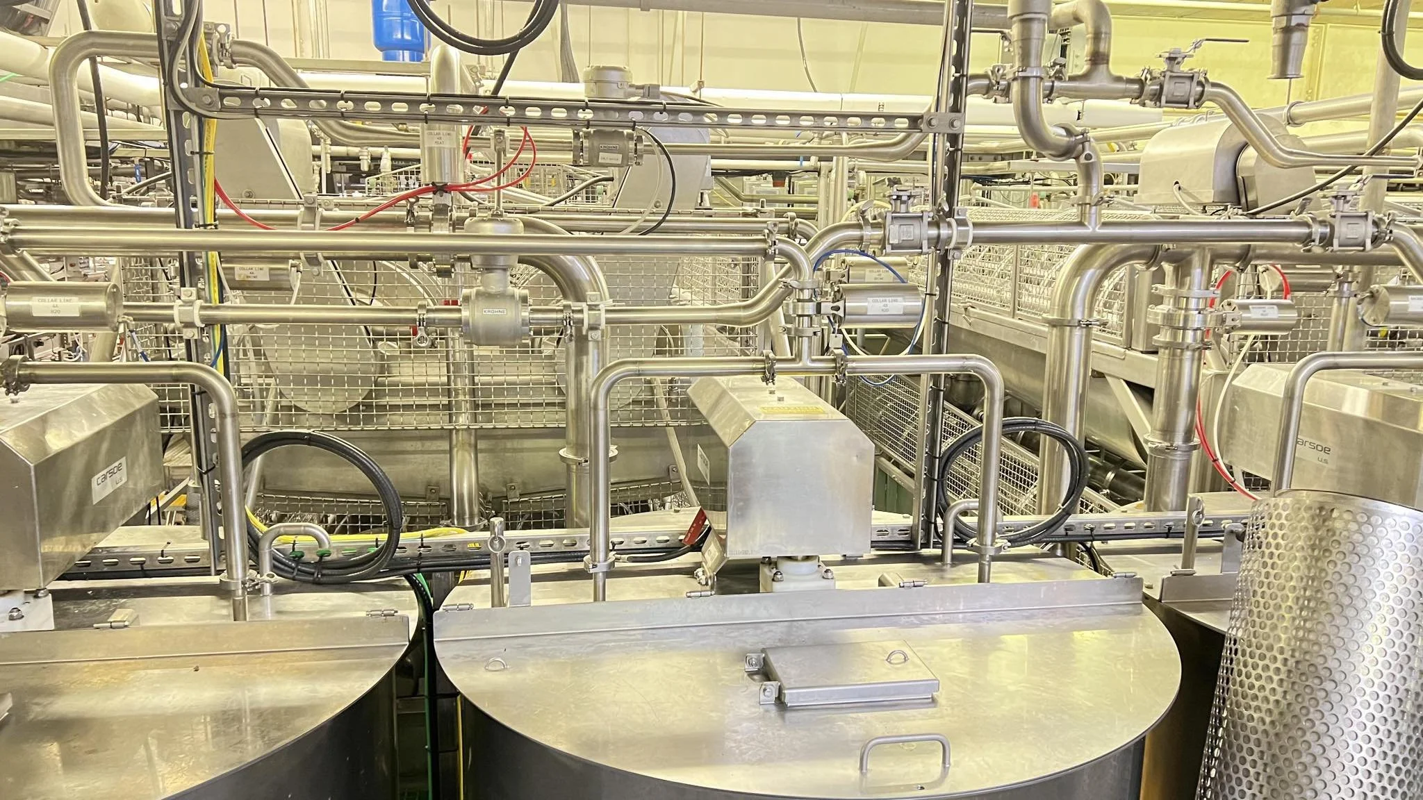 Close-up view of stainless steel piping system with valves, gauges, and electrical wiring in industrial setting.