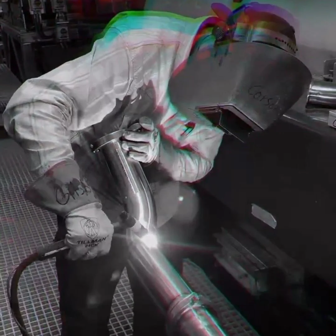 Worker welding a metal pipe in a workshop, wearing protective gloves and a welding helmet.