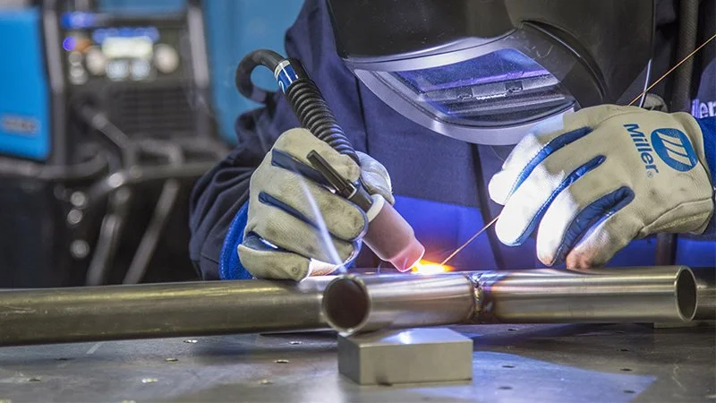 A person wearing a welding helmet, gloves, and dark clothing welds a metal pipe with a welding torch, creating sparks in a workshop.