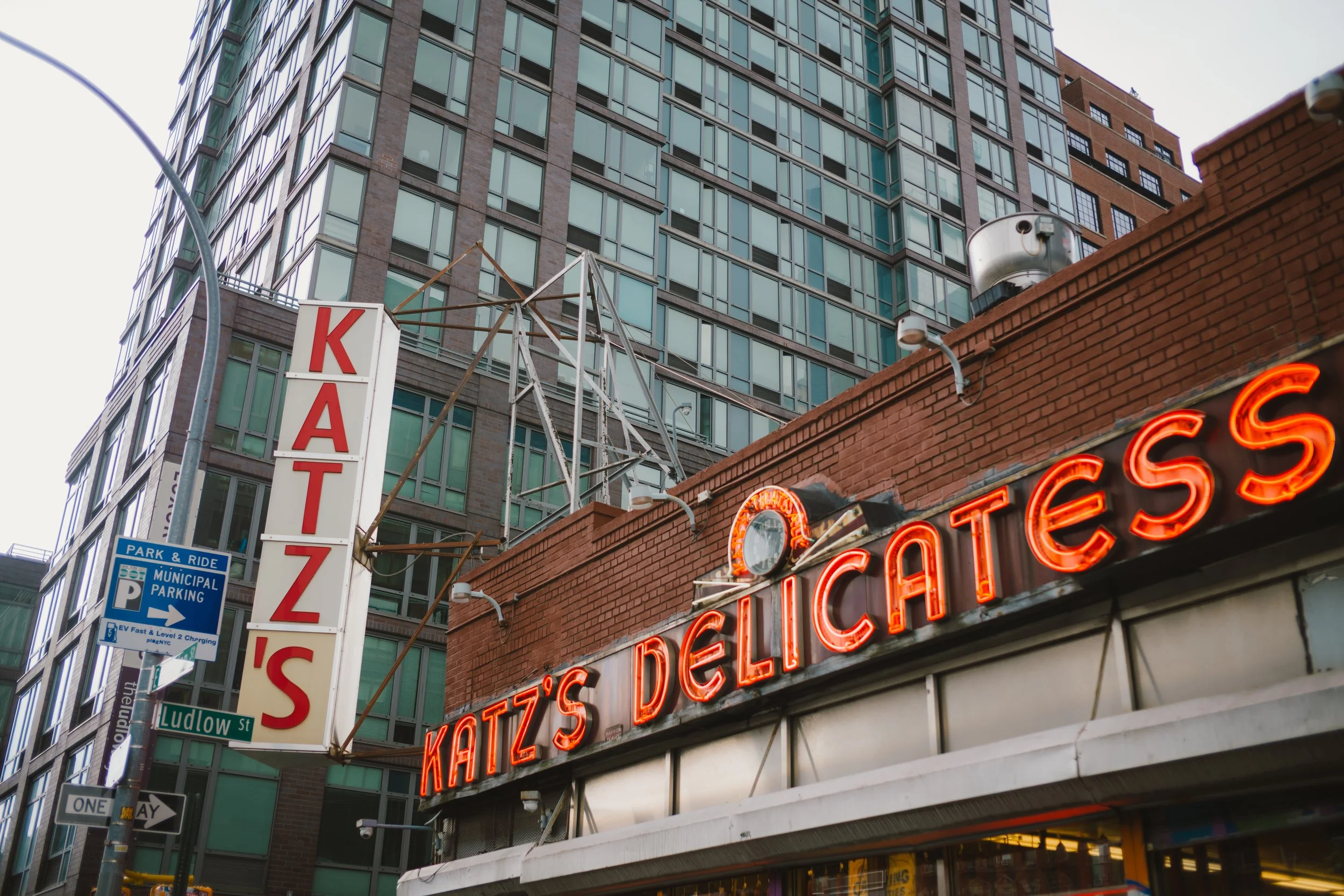 Street view featuring a karaoke sign and a neon sign for Katz's Delicatessen, with tall buildings in the background and several street signs including parking and one-way indications.